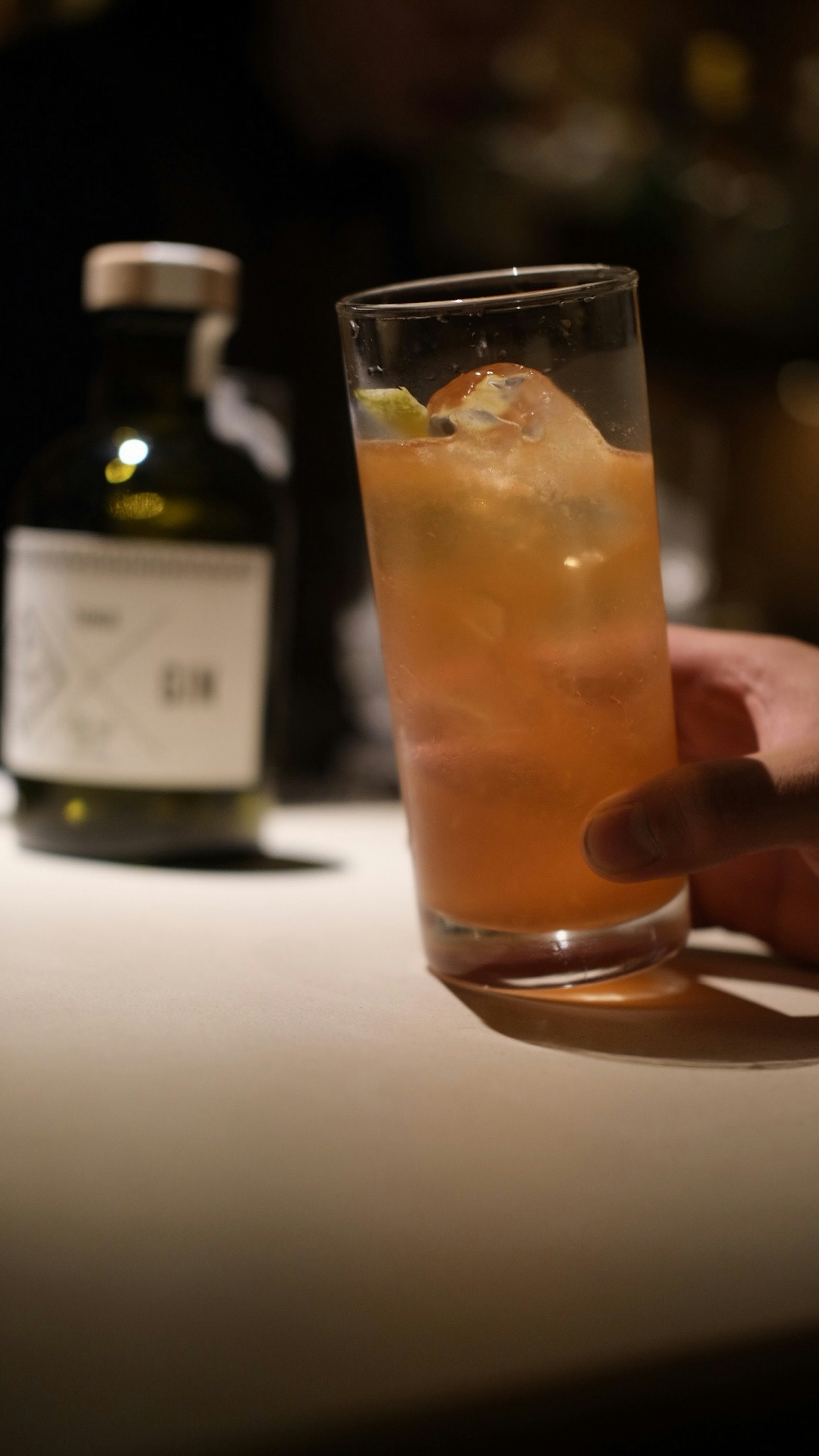 A hand holding a cocktail glass with ice and a bottle in the background