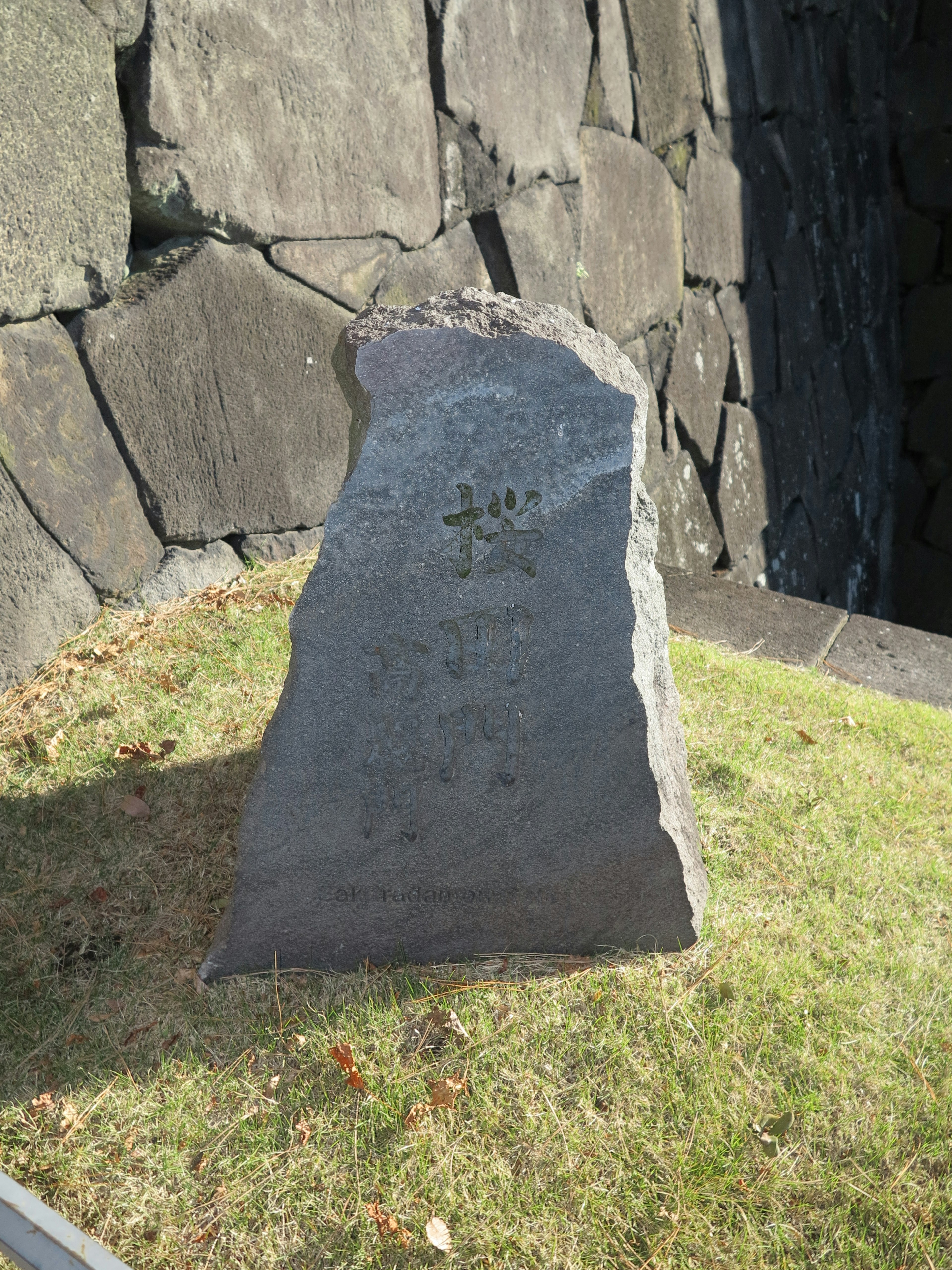 Ein Steindenkmal steht auf Gras mit einer Steinmauer im Hintergrund und Inschriften auf dem Stein