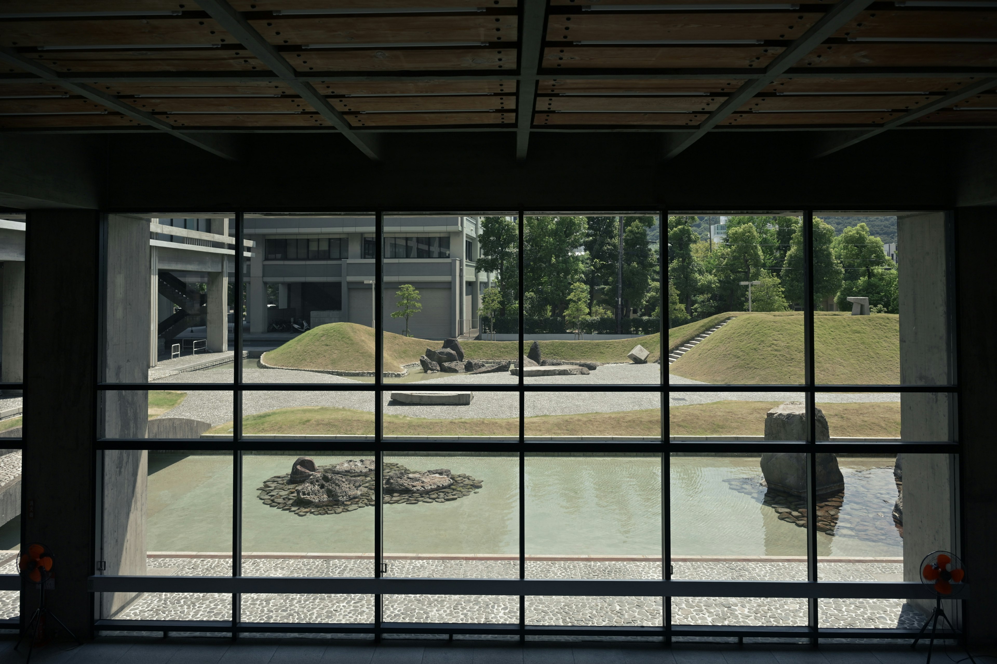 Vue d'un jardin japonais à travers une fenêtre d'un bâtiment moderne avec des collines vertes et des rochers près d'un étang tranquille