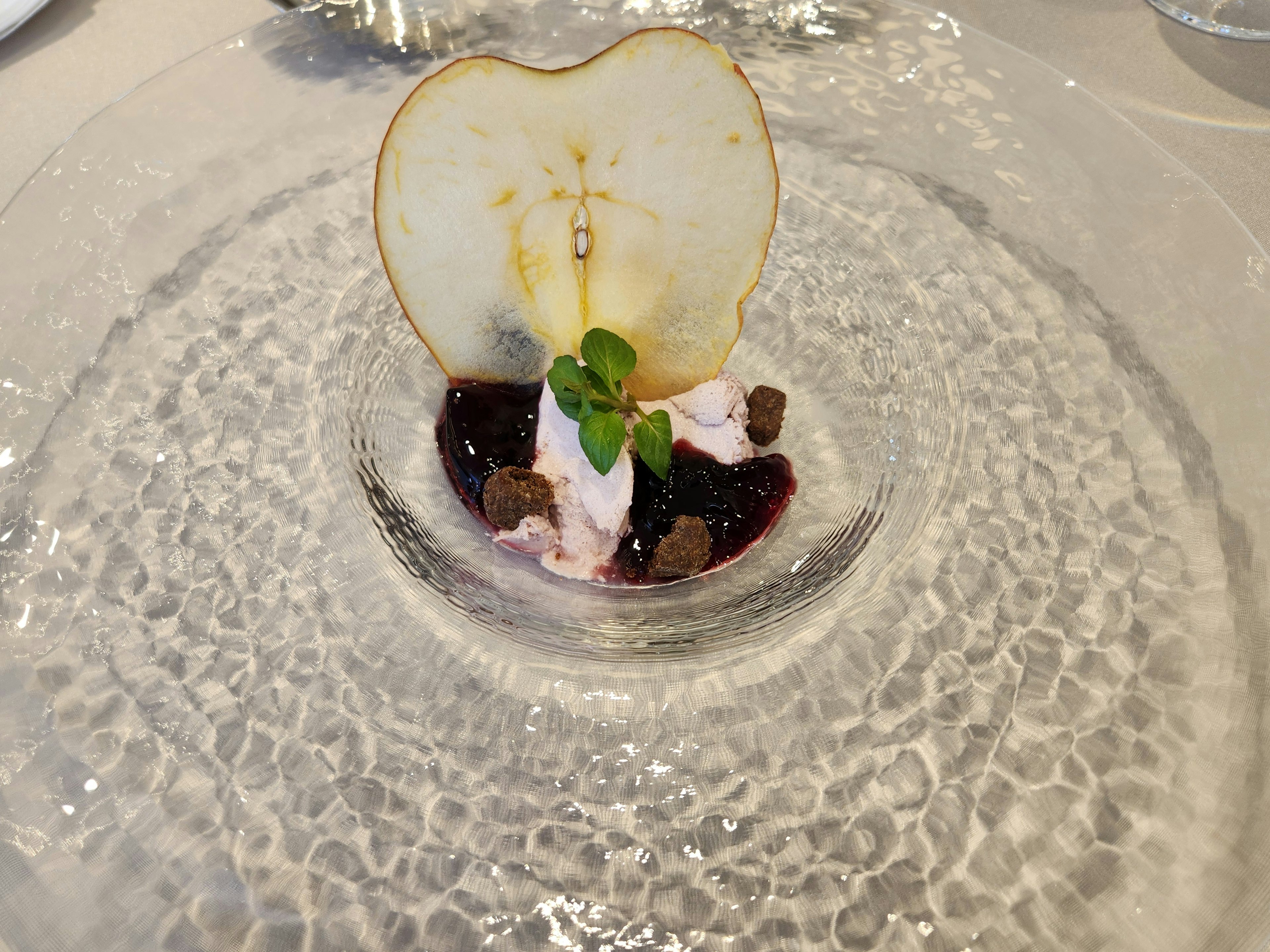 Dessert featuring a heart-shaped apple slice on a clear plate with purple sauce and mint