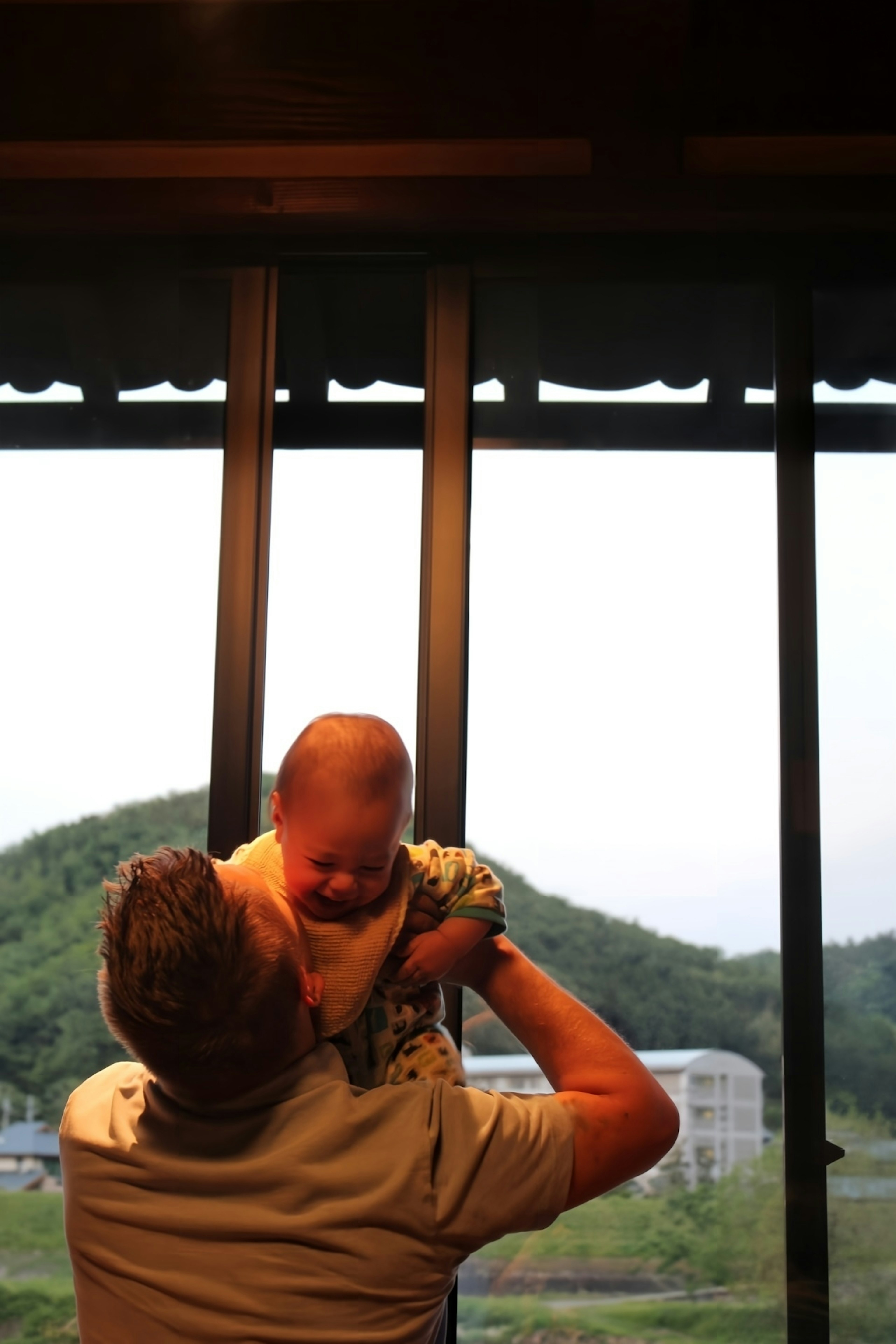 Ein Vater hebt ein Baby mit Blick auf grüne Hügel durch das Fenster