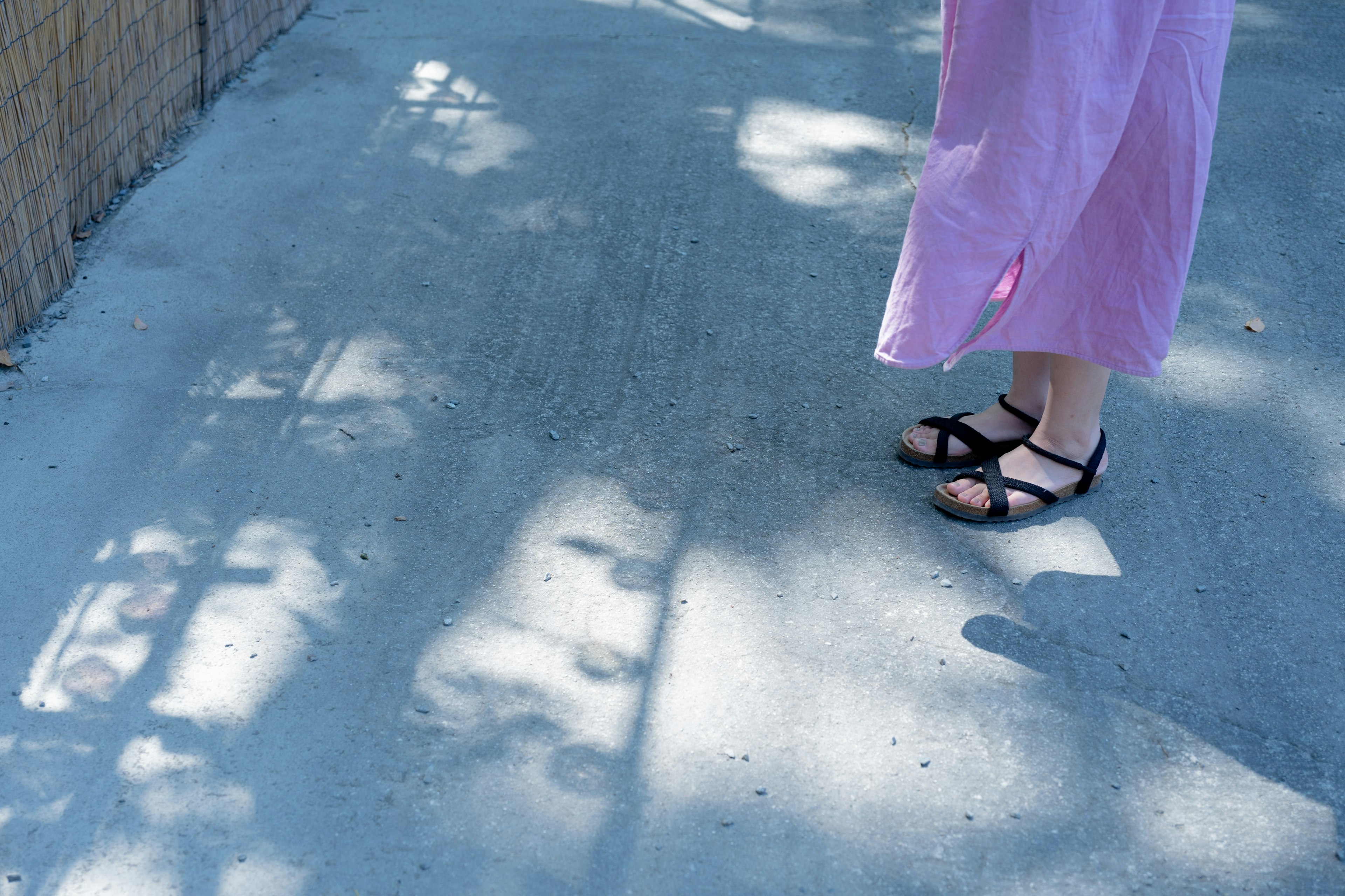 Feet in black sandals and pink dress standing on a sunlit concrete path with shadows