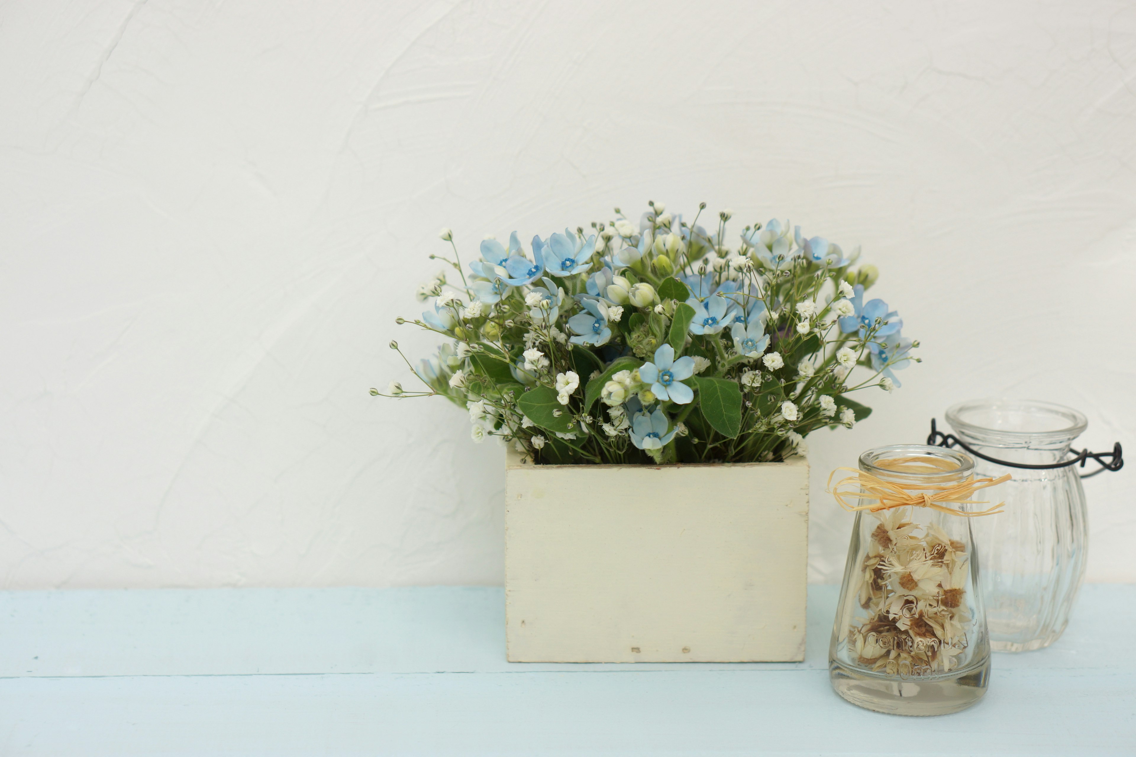 A flower pot with blue flowers and white blooms alongside glass jars on a light blue surface