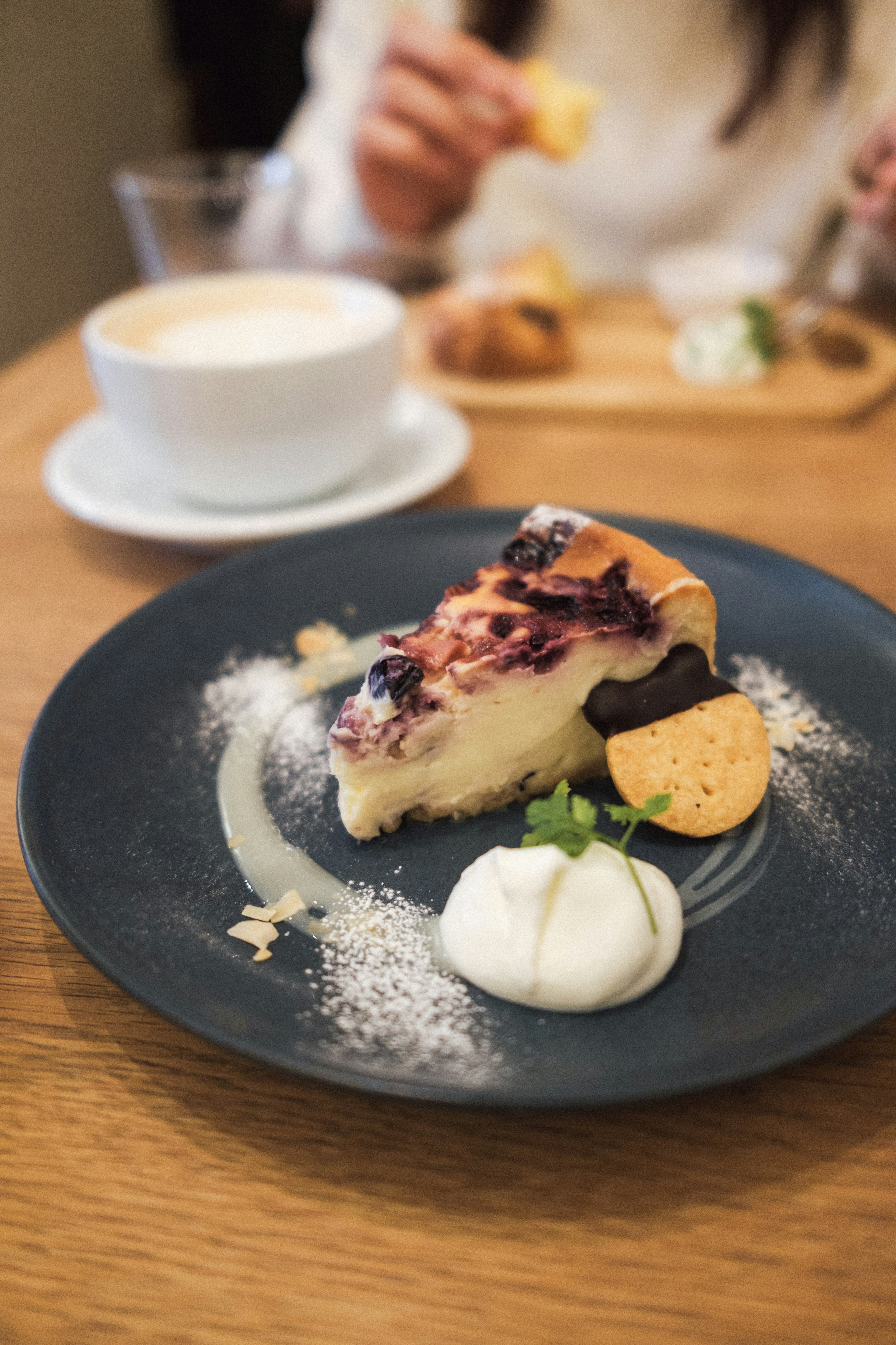 Ein Stück Blaubeerkäsekuchen auf einem dunklen Teller mit Sahne und einem Keks Kaffee im Hintergrund