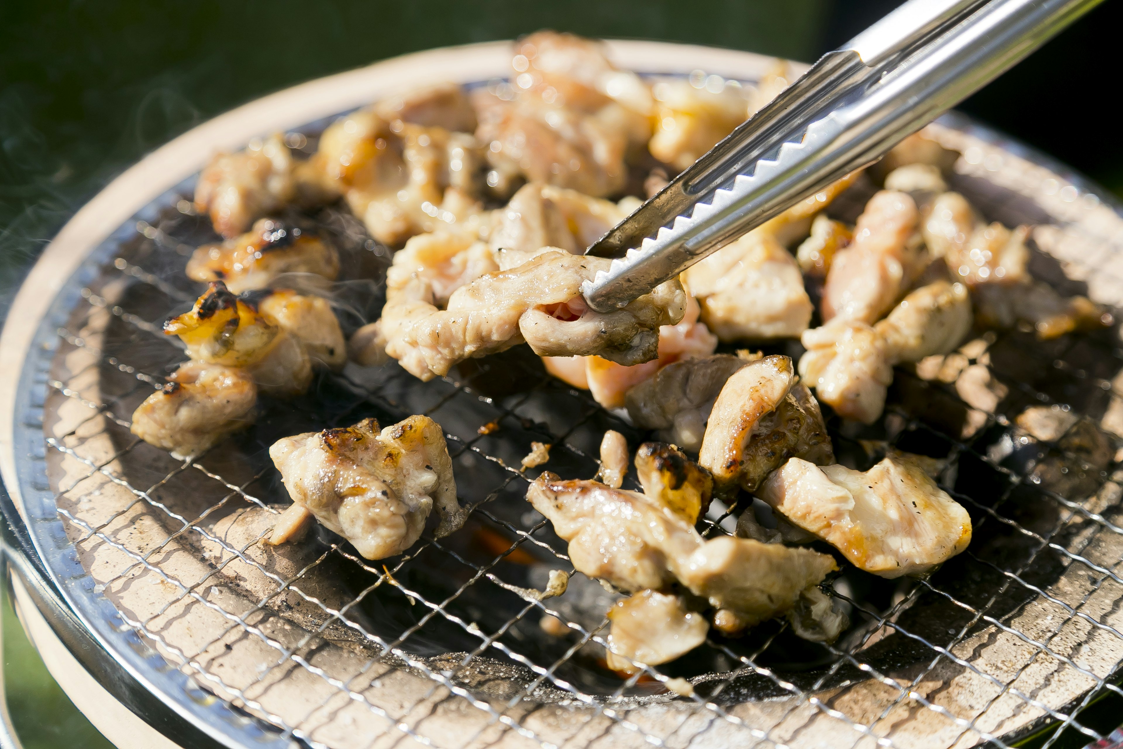 焼かれている鶏肉の塊が網の上にあり、トングで持ち上げられている