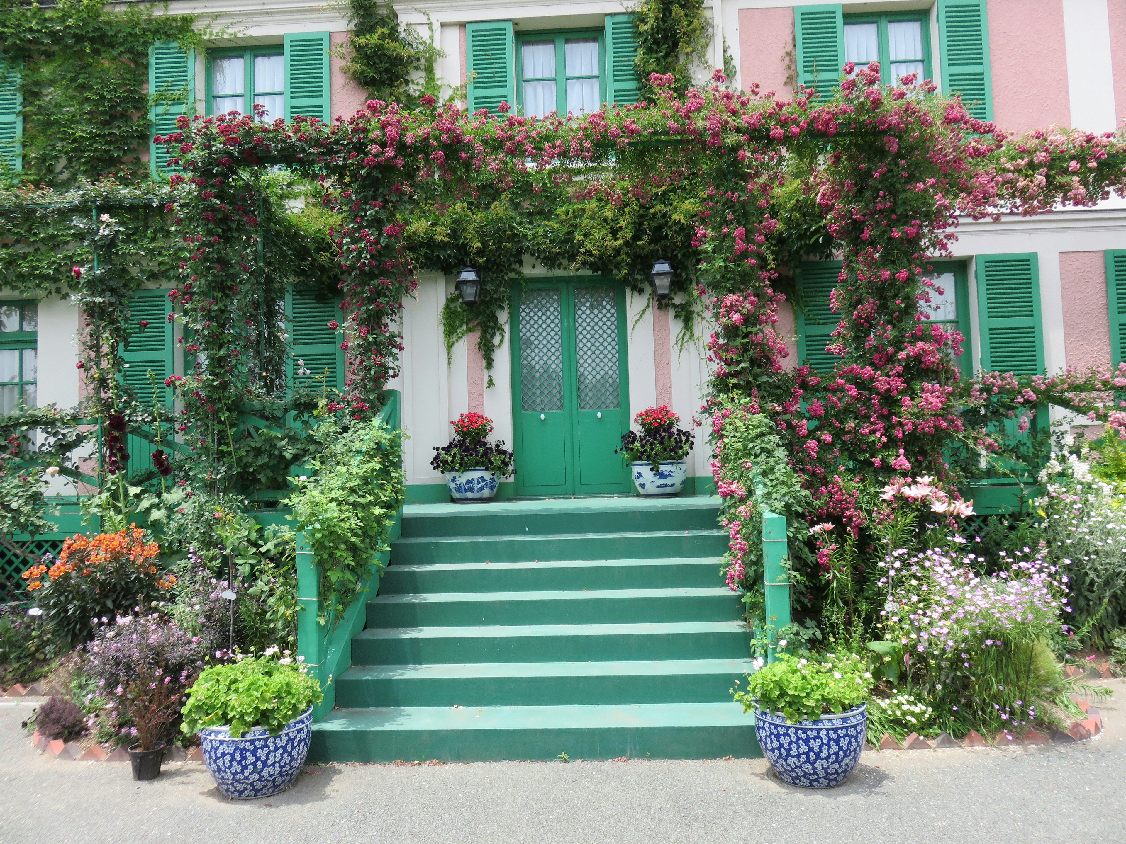 Pink house with green doors and windows adorned with lush plants and flowers