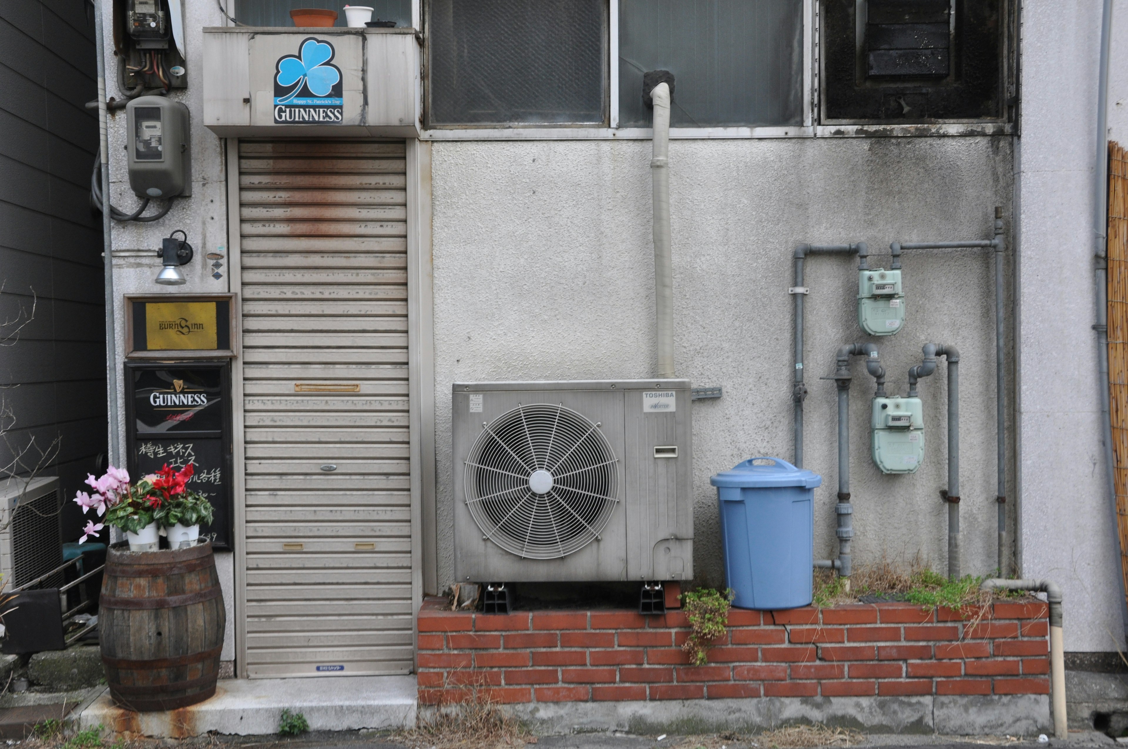 Exterior de un edificio antiguo con una unidad de aire acondicionado y un bote de basura azul decorado con un barril de flores