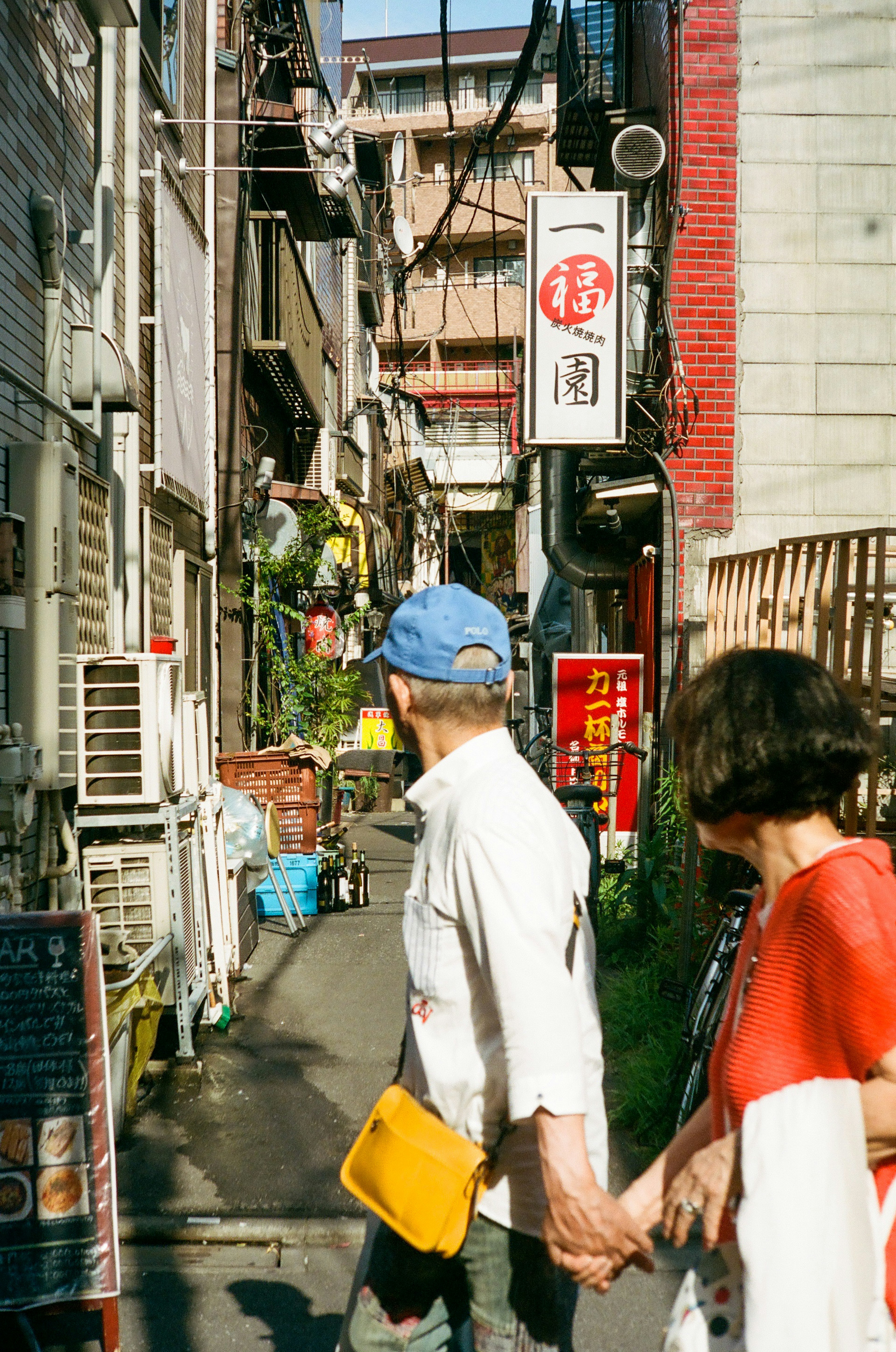 狭い路地を歩くカップルが手をつないでいる様子で周囲には飲食店の看板が見える