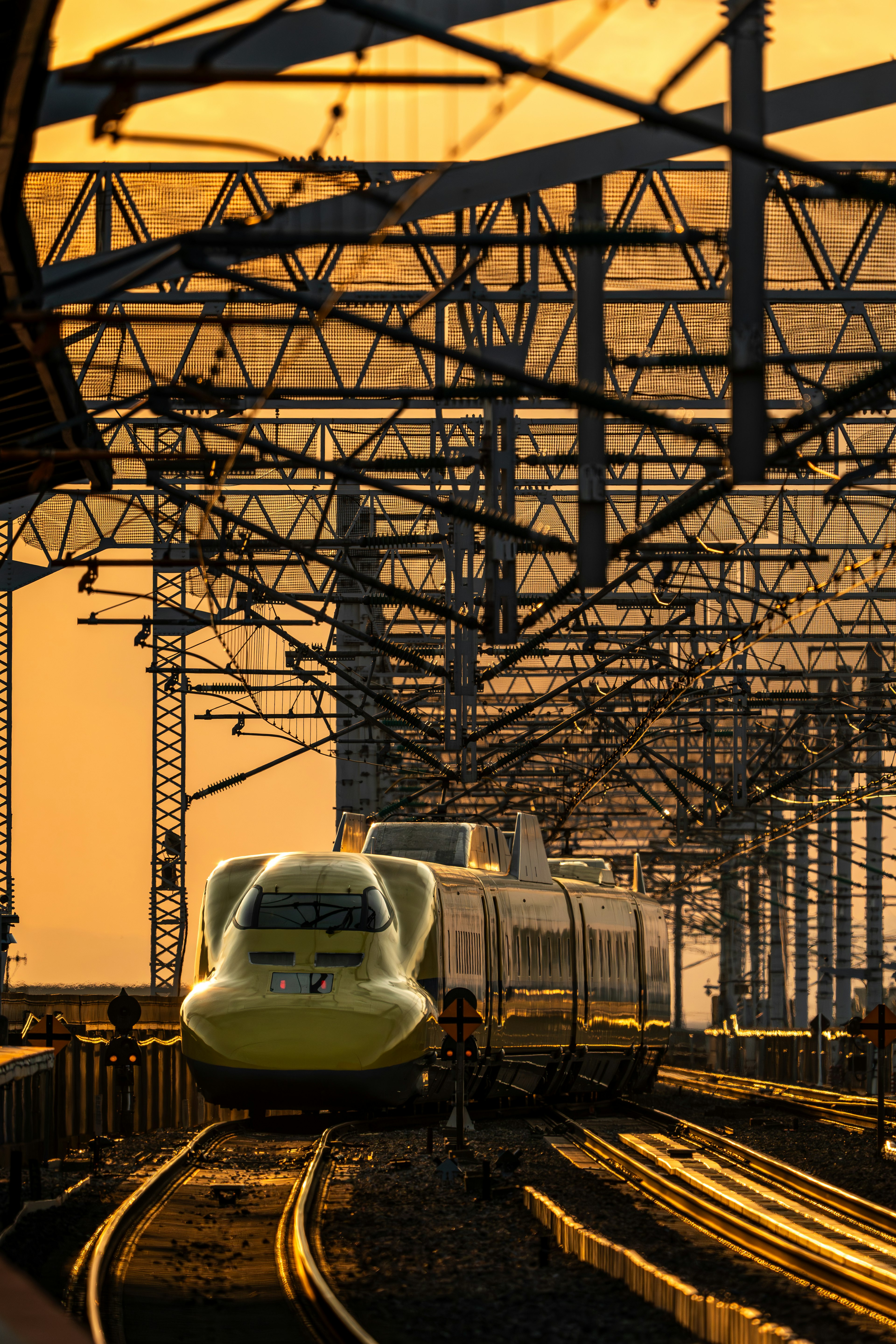 Yellow Shinkansen train in sunset with railway overhead structures