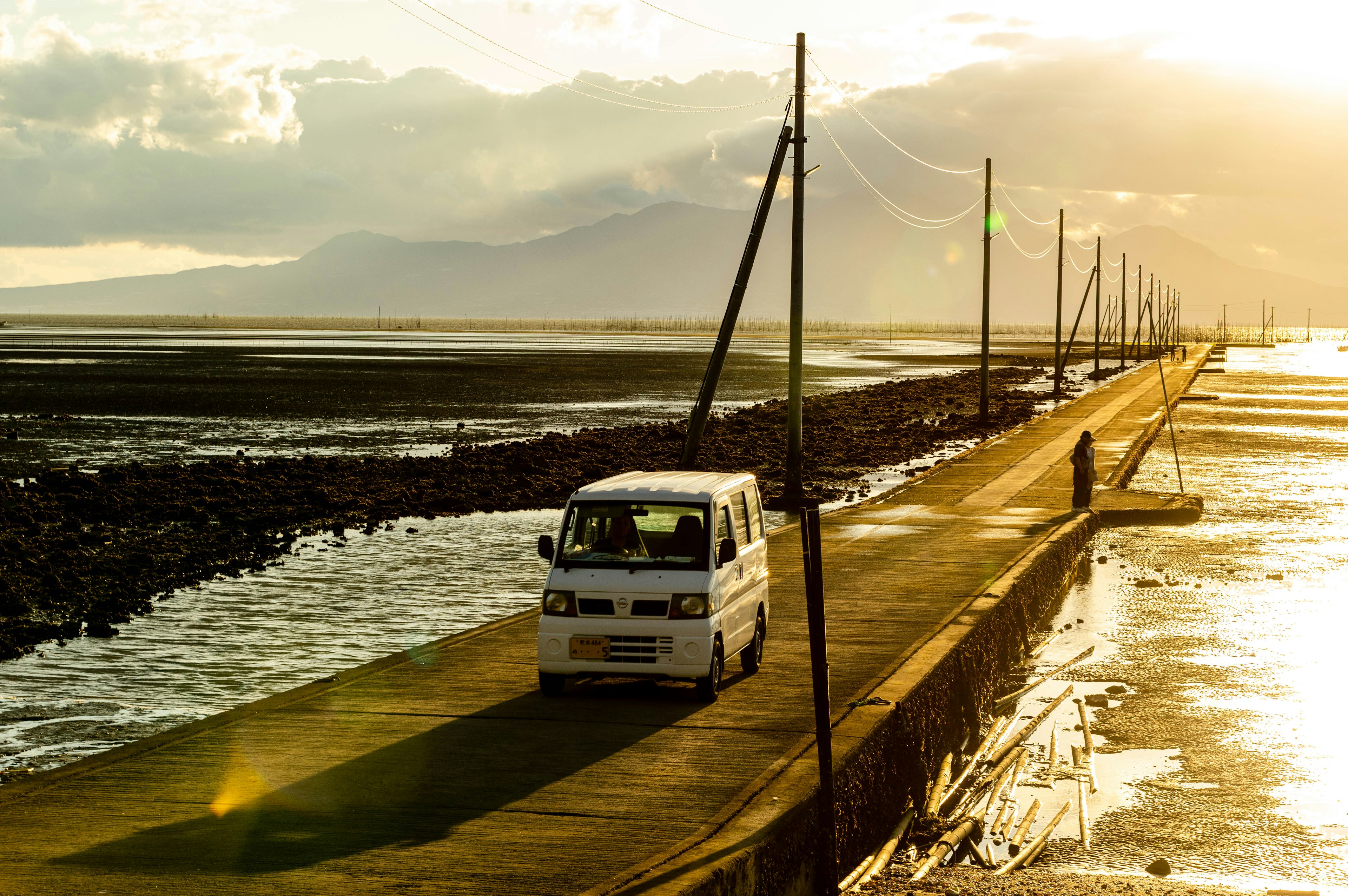 Ein weißer Van fährt auf einer Küstenstraße, umgeben von Wasser und Sonnenuntergang