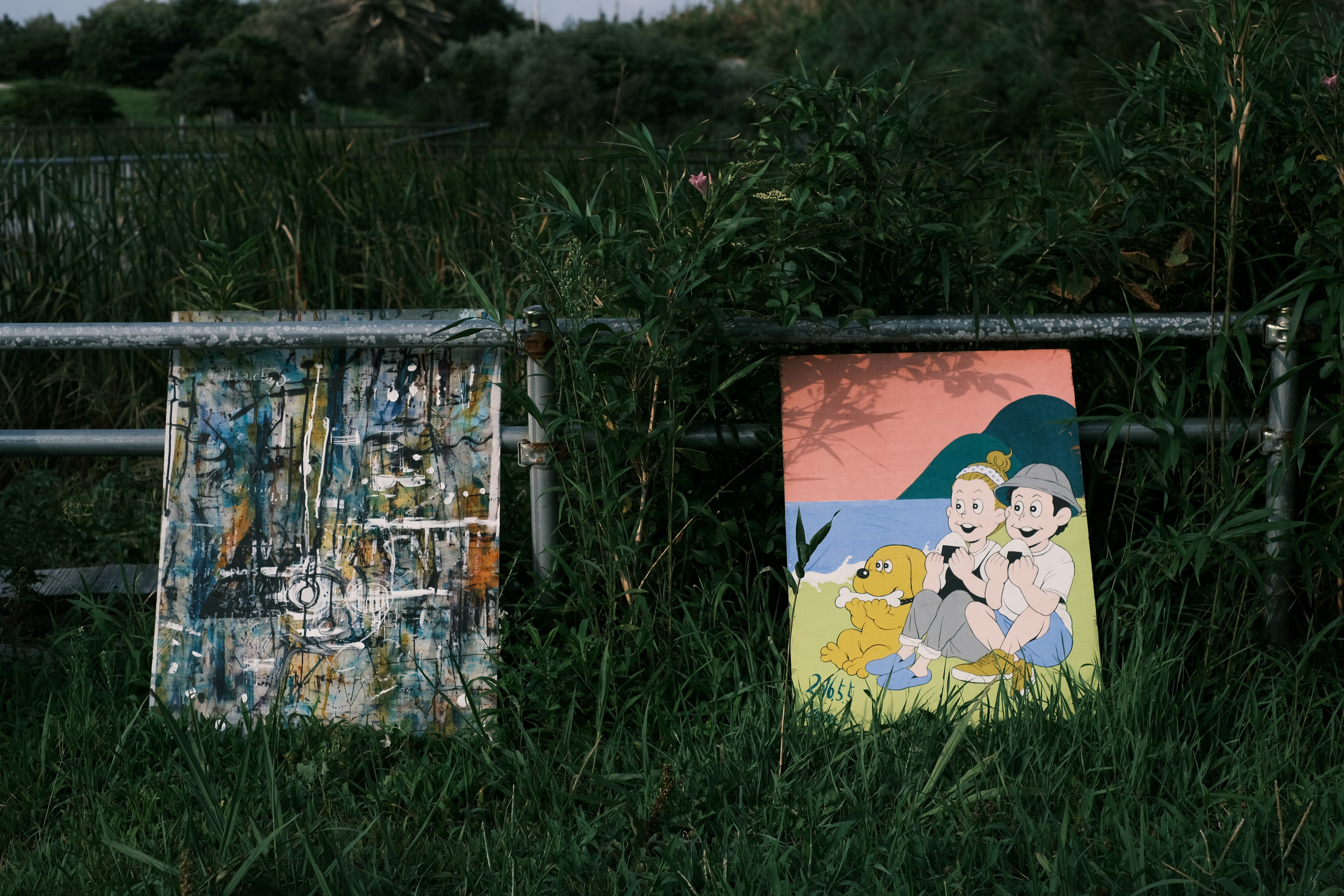 Deux peintures appuyées dans l'herbe verte l'une est une œuvre abstraite colorée l'autre présente des personnages dans un paysage