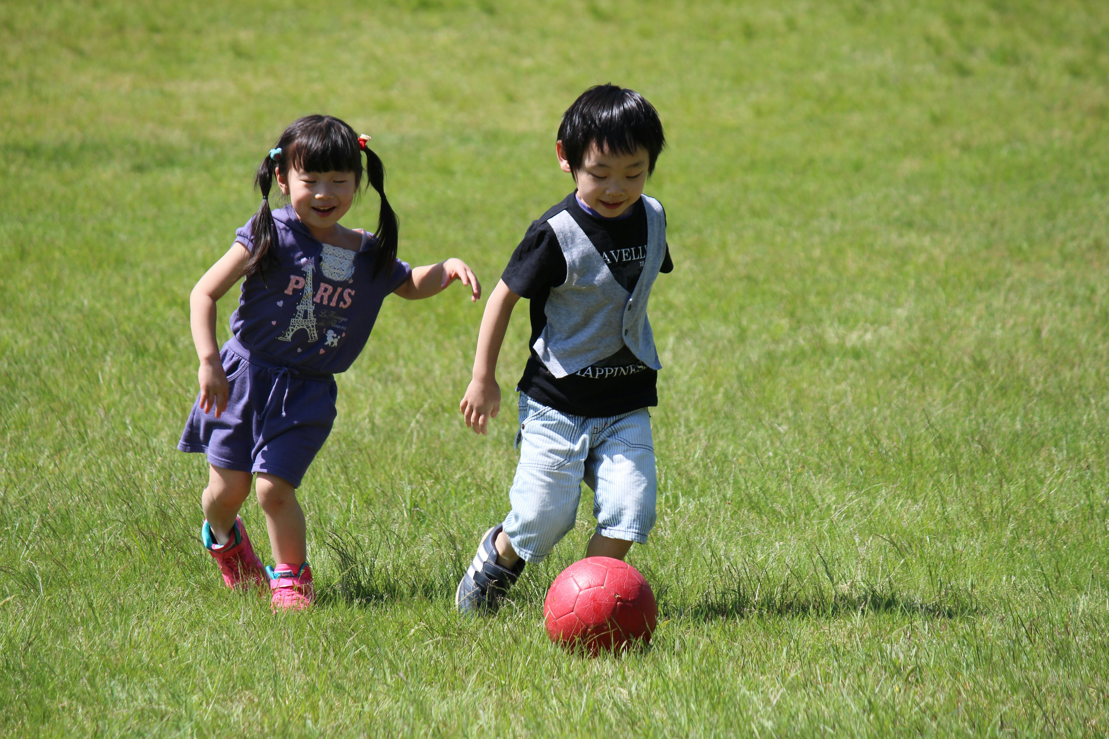 Zwei Kinder, die auf dem Gras einem roten Ball nachjagen