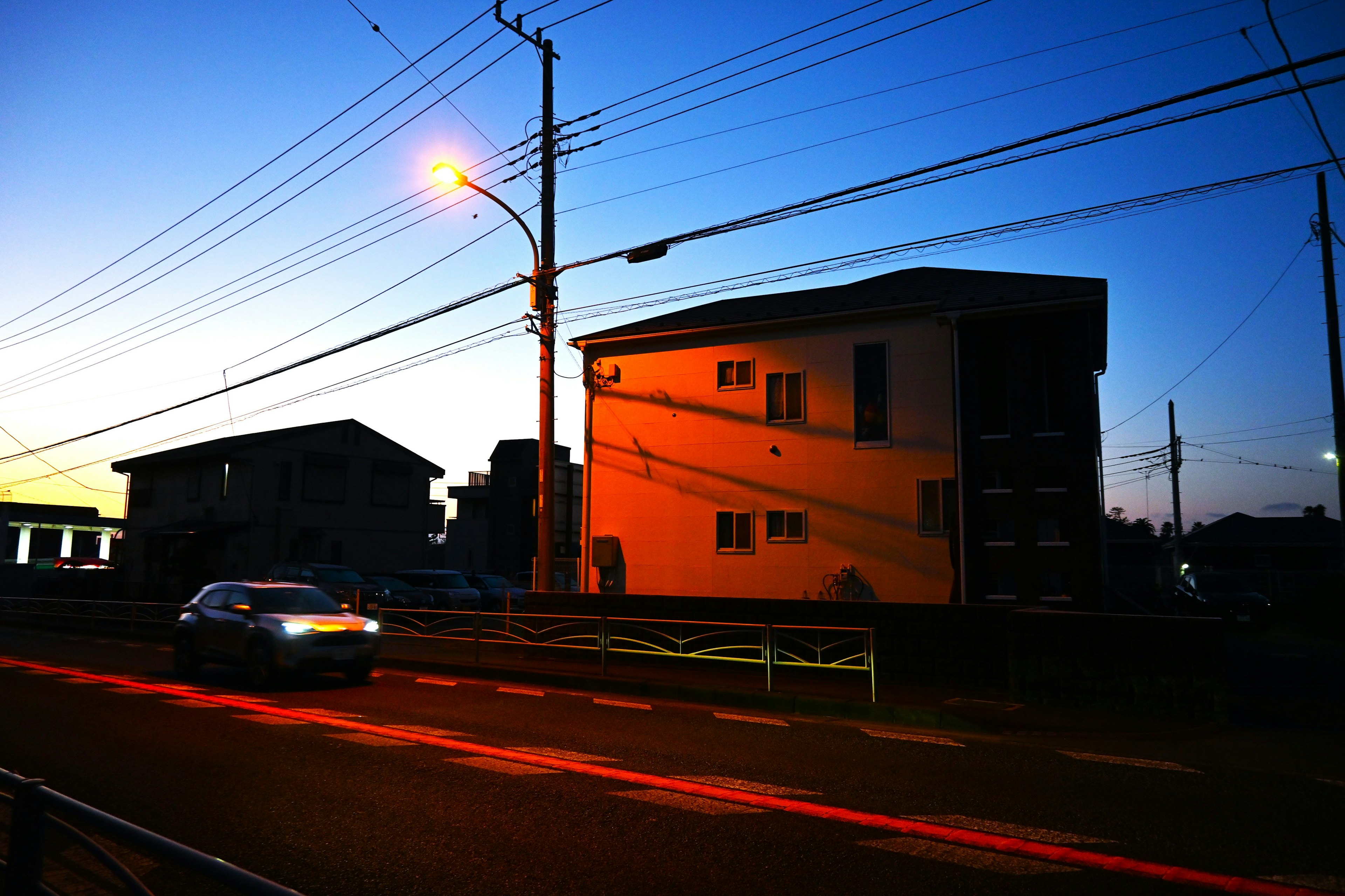 Paisaje urbano al atardecer con un coche pasando y casas iluminadas