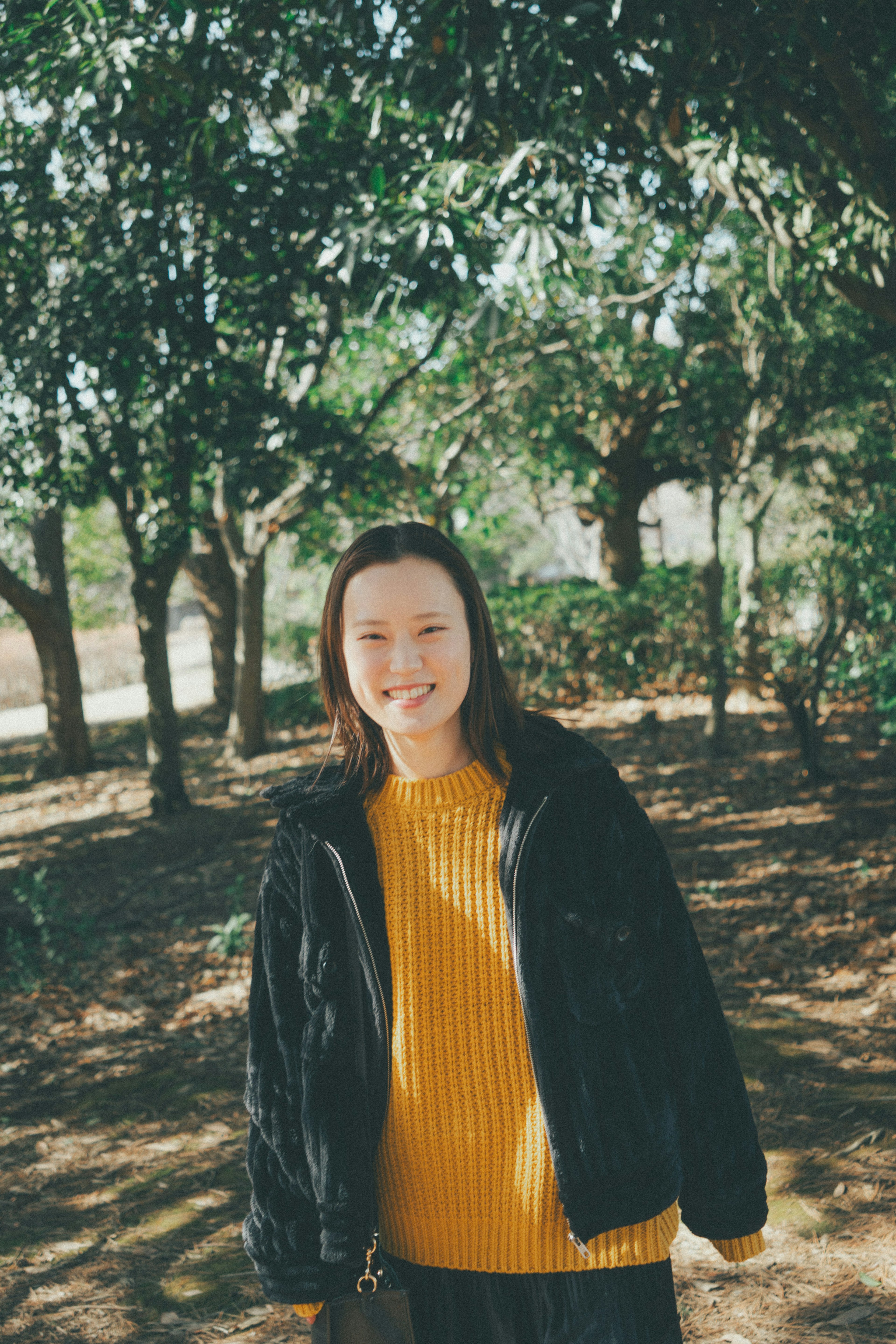 Une femme souriante se tenant parmi des arbres verts portant une veste noire et un pull jaune