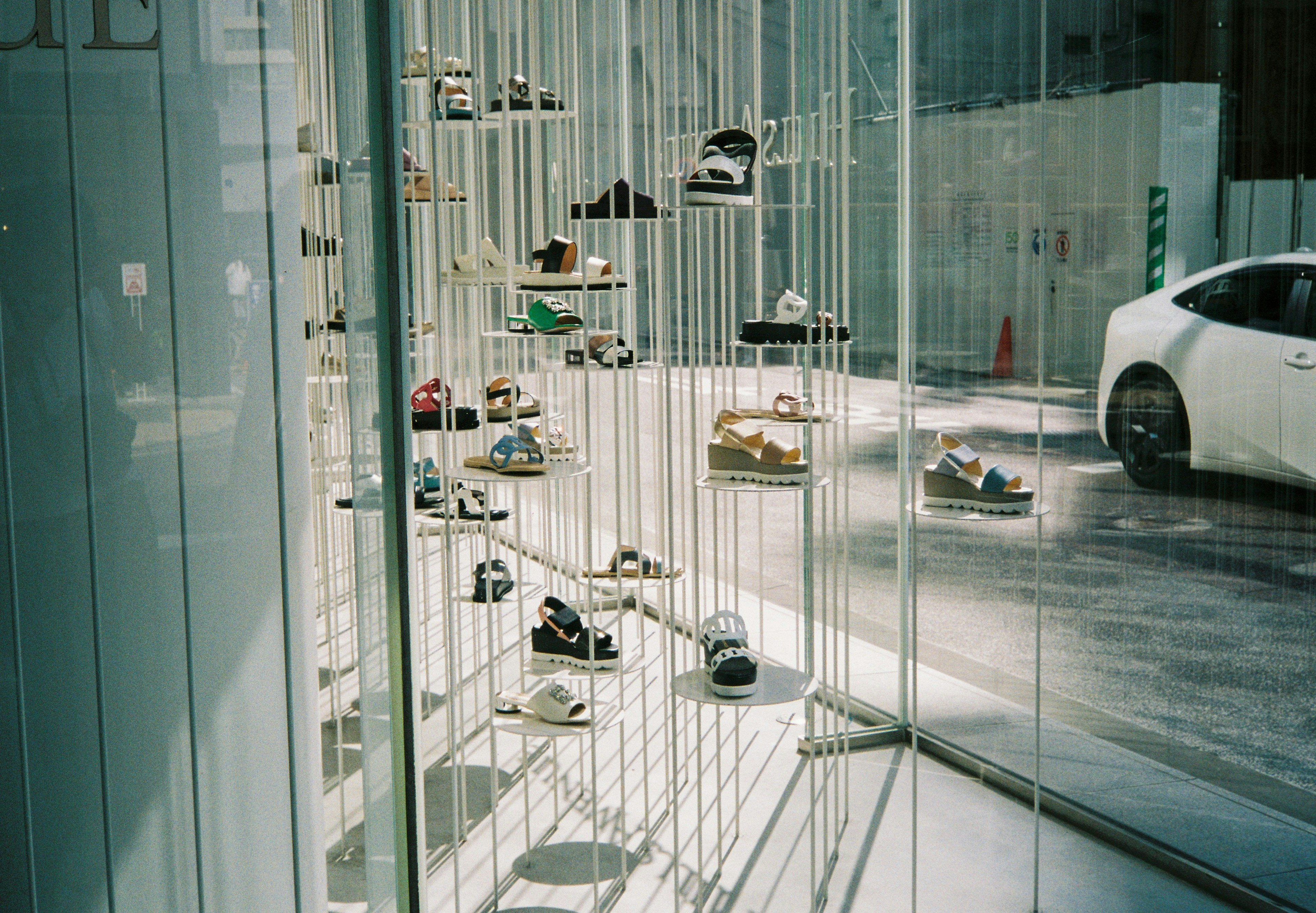 Various shoes displayed in a transparent storefront with a white car outside