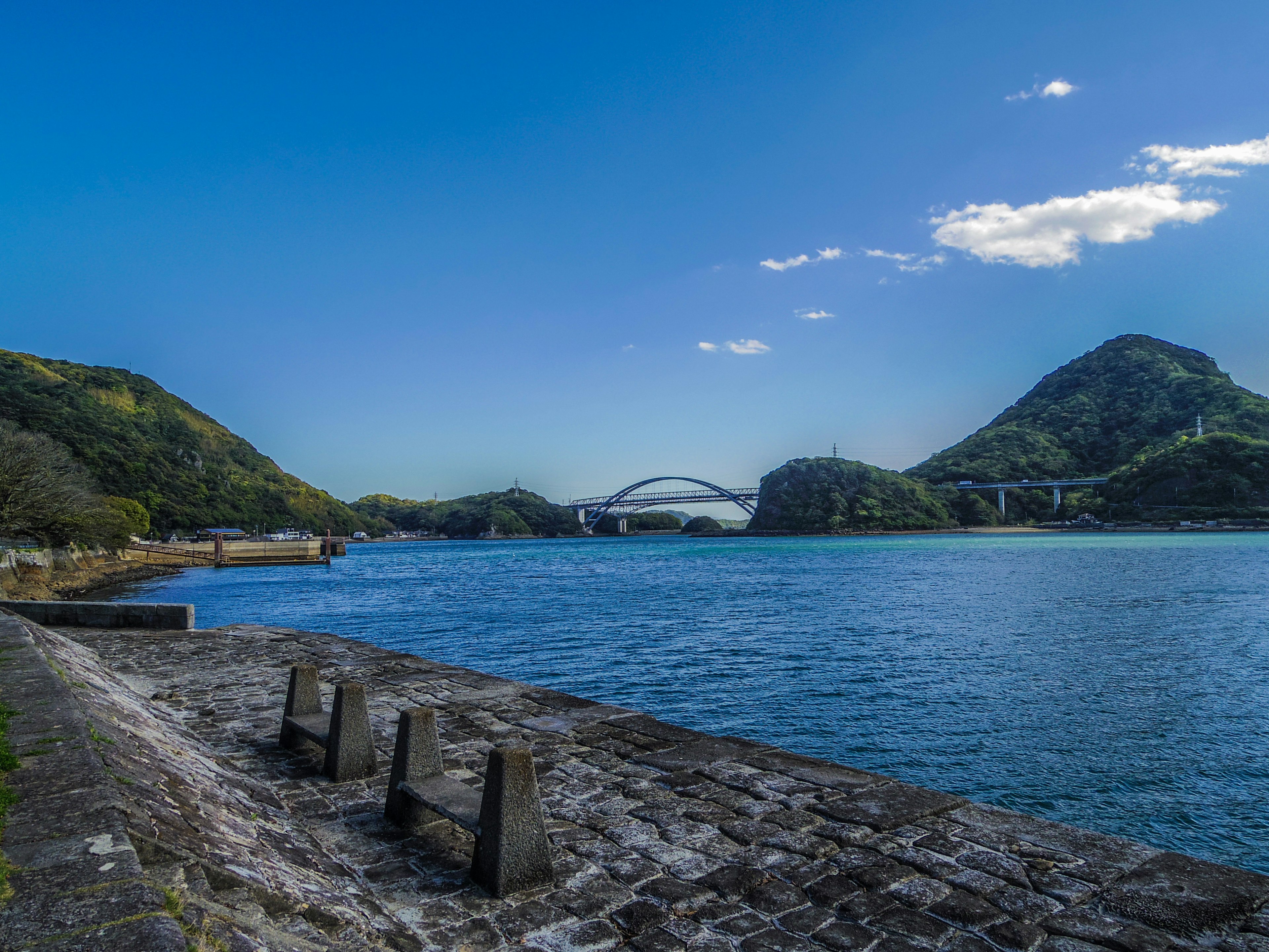 Paisaje marino sereno con cielo azul y montañas de fondo con un puente a lo lejos