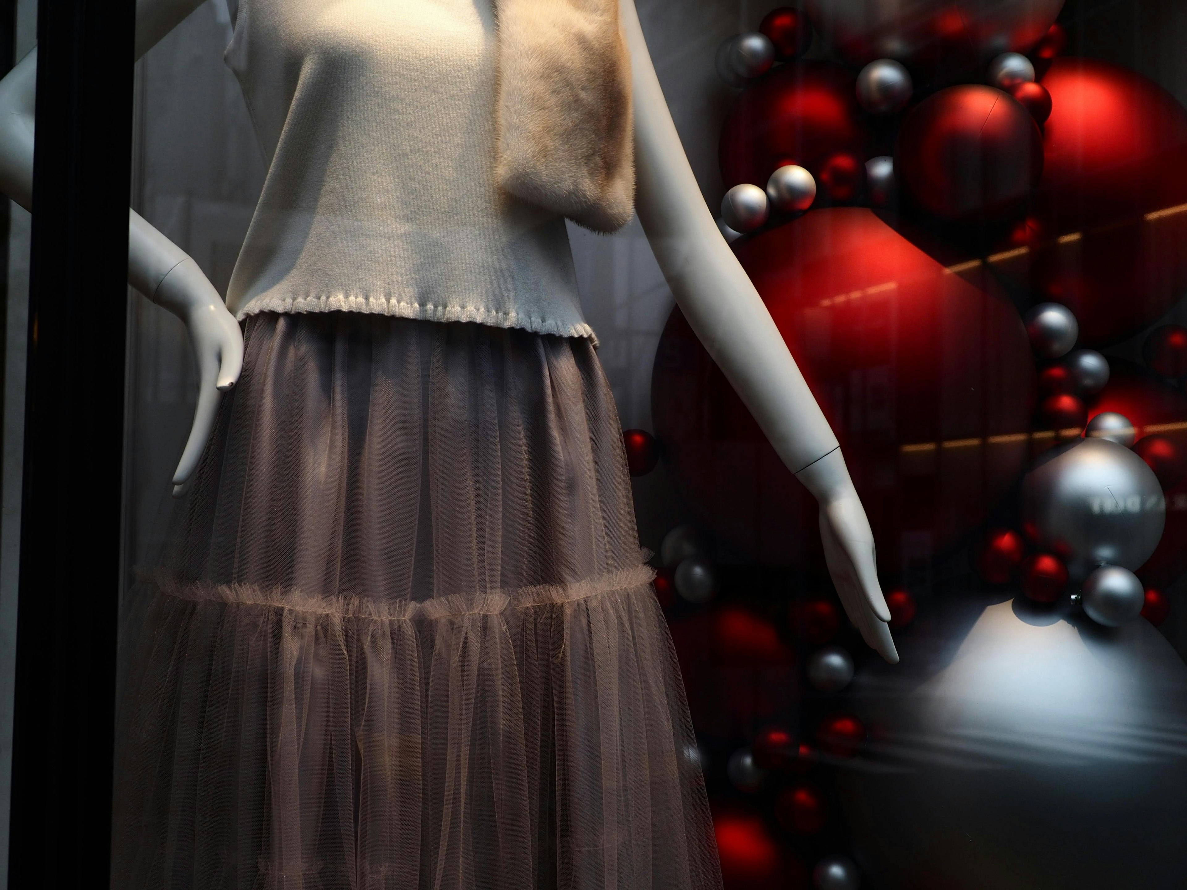 A mannequin in a window display wearing a white top and gray skirt with a background of red and silver decorations