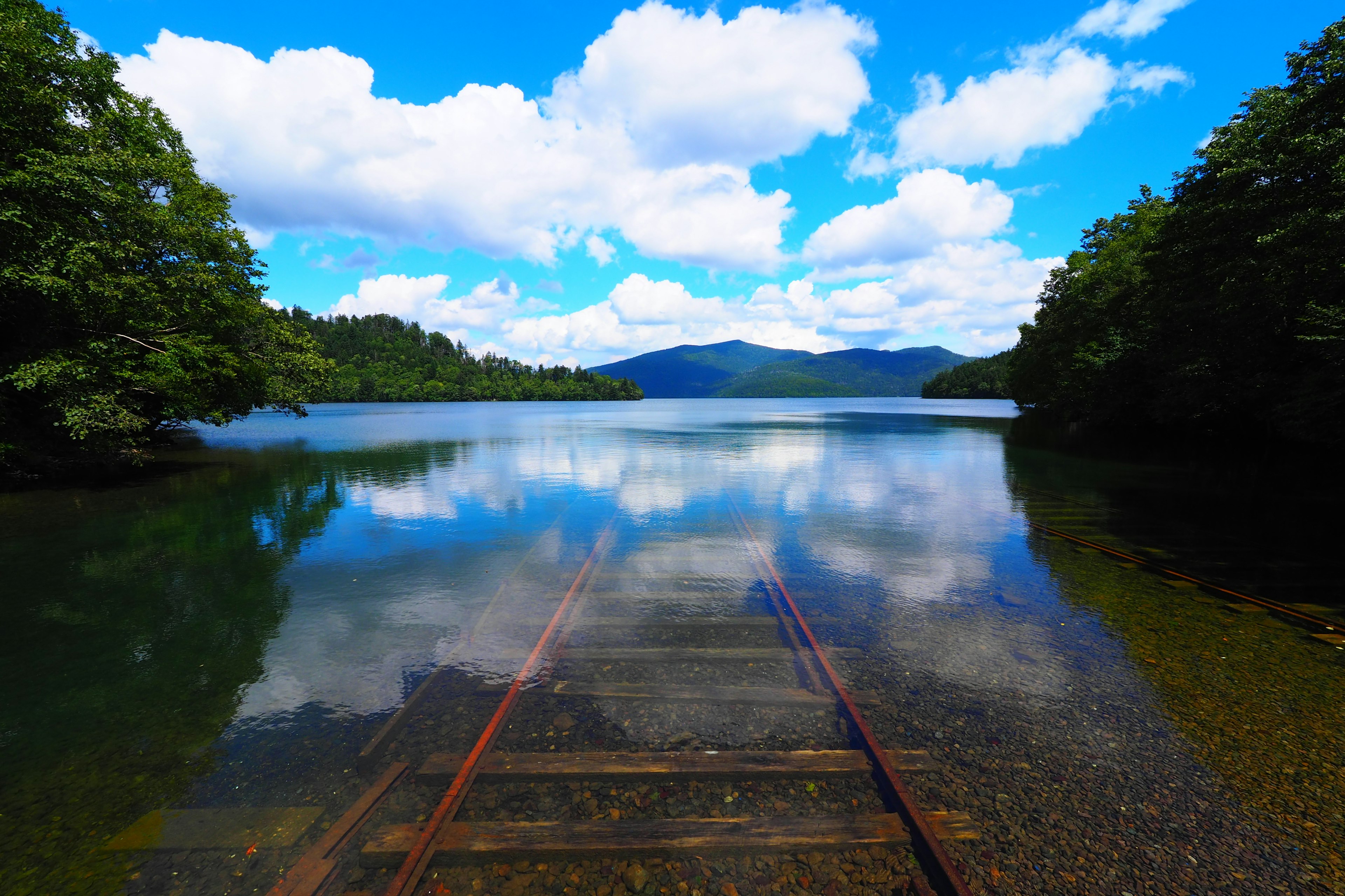 Paisaje de lago sereno con cielo azul y nubes blancas reflejadas Vías de tren oxidadas que se extienden en el agua