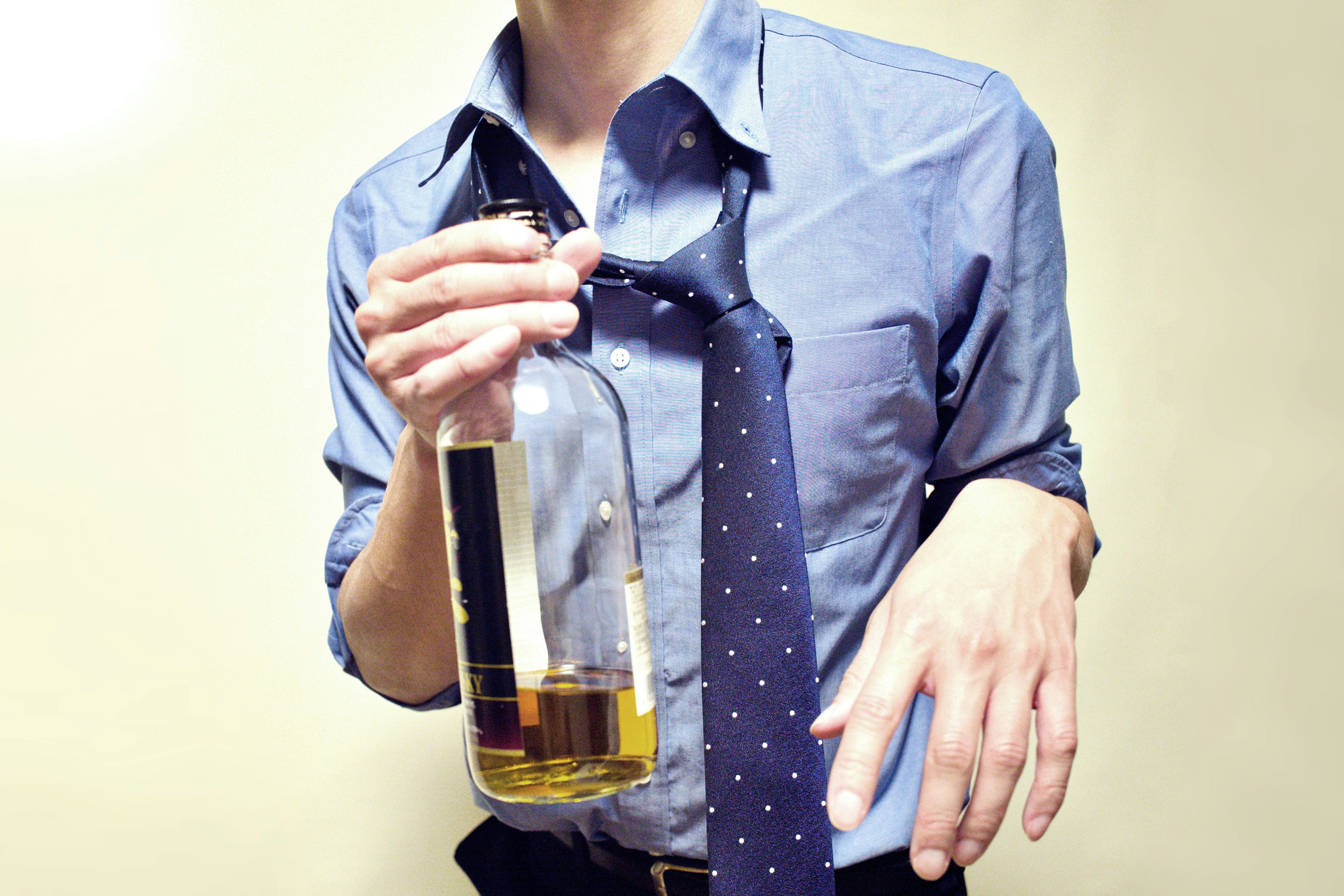 A man in a dress shirt holding a bottle of liquor with a tie loosely around his neck