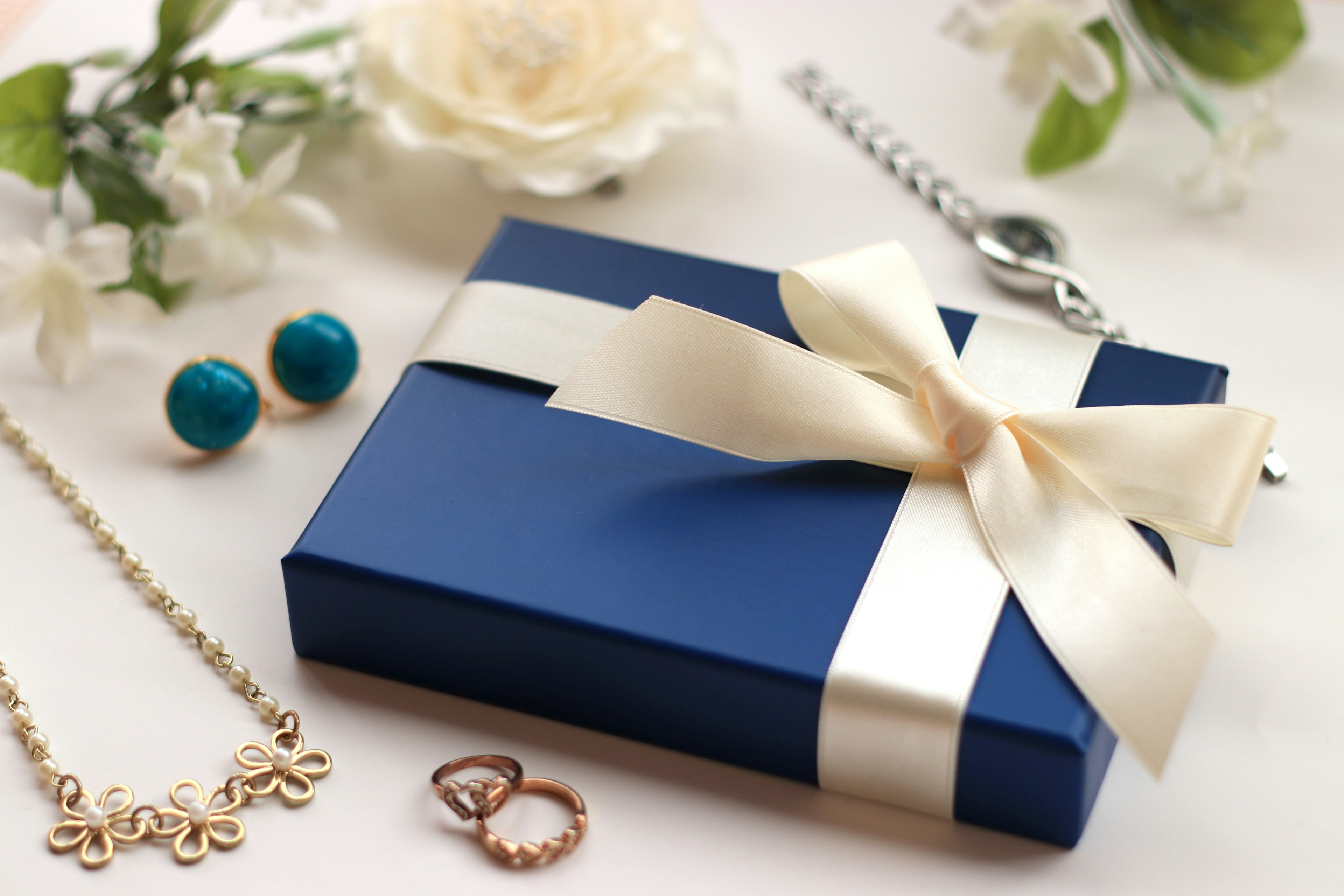 A blue gift box with a white ribbon surrounded by jewelry and flowers