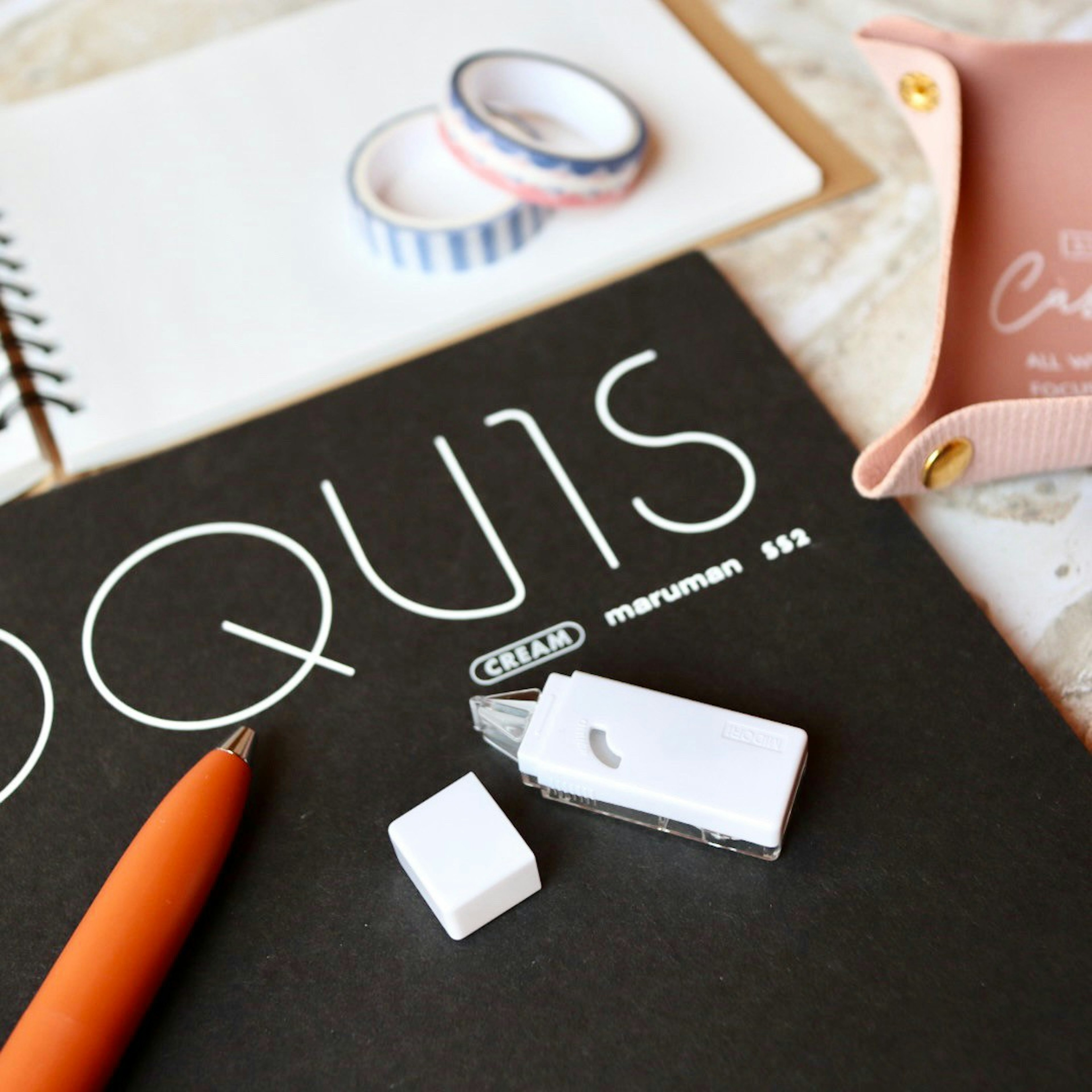 Black notebook with white text orange pen white eraser white cutter colorful washi tape and pink pencil case on a desk