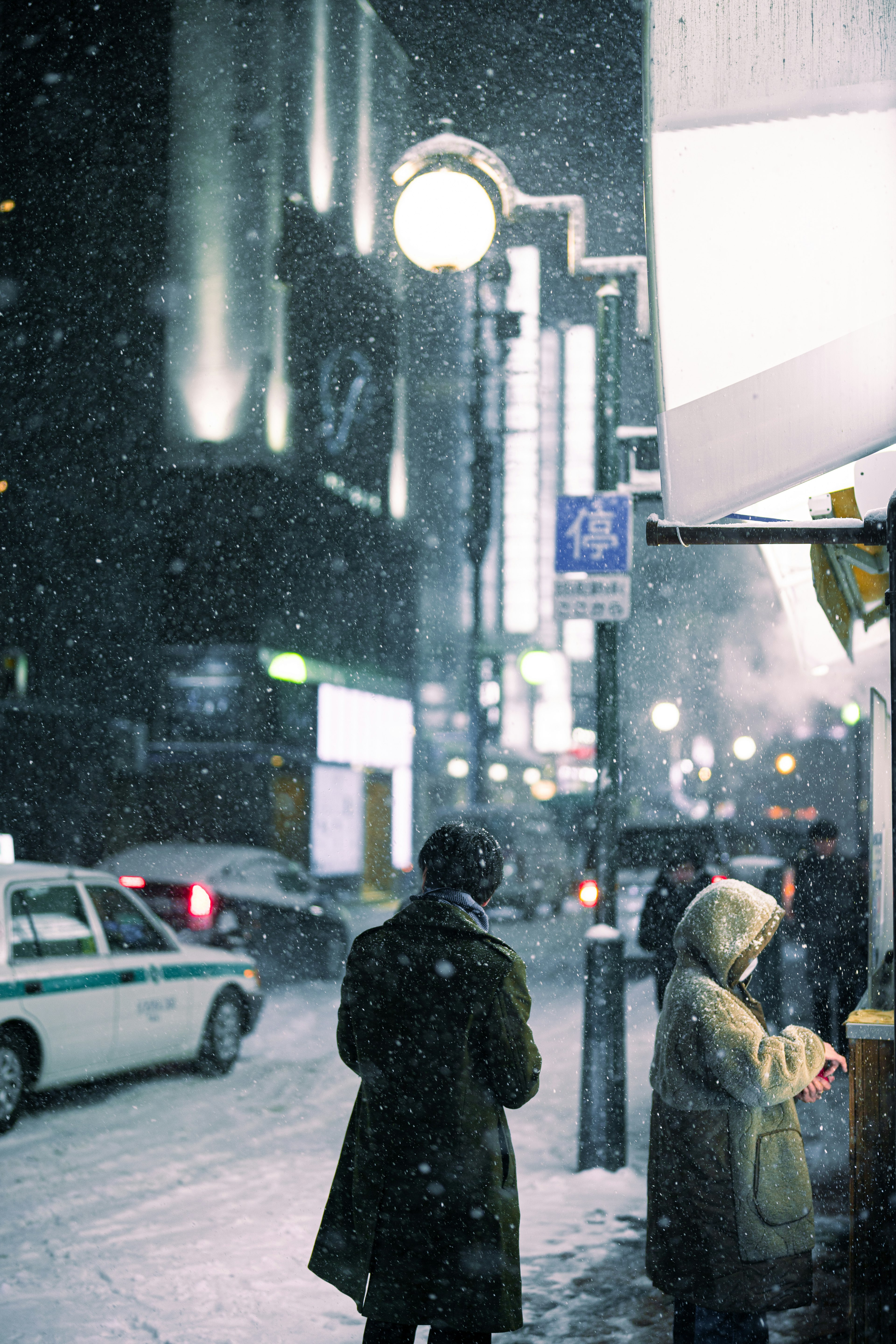 Two figures standing on a snowy street corner at night with taxi lights