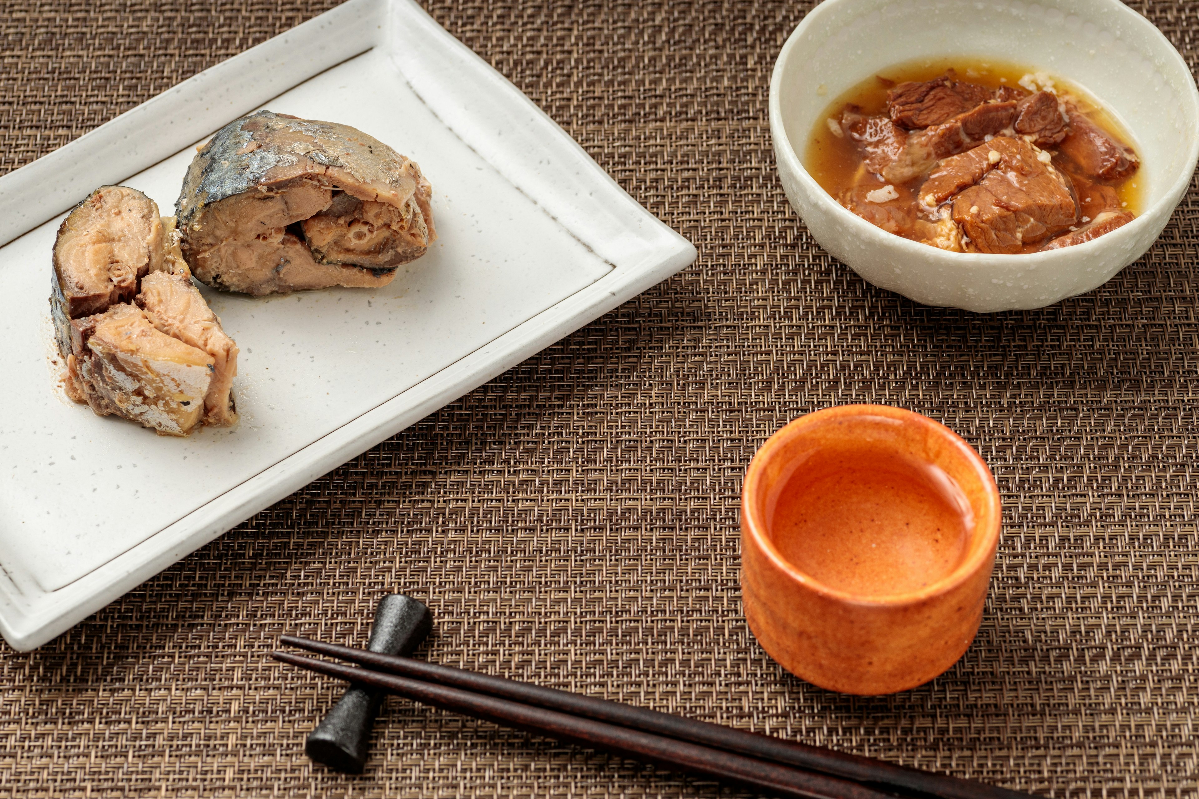 A plate with fish pieces and a bowl of sauce alongside chopsticks and a small cup