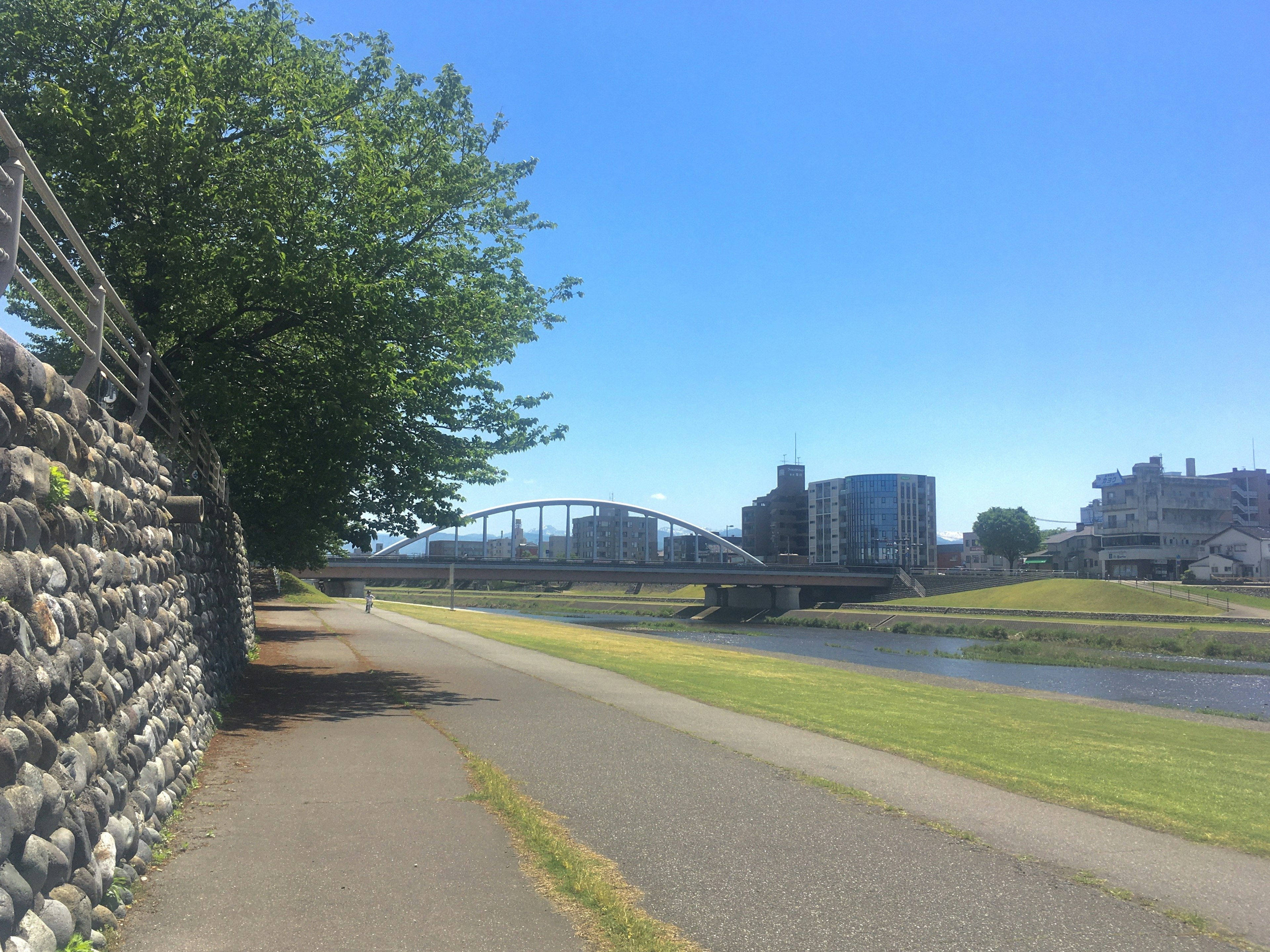 Walking path along a river with a bridge and buildings under a blue sky