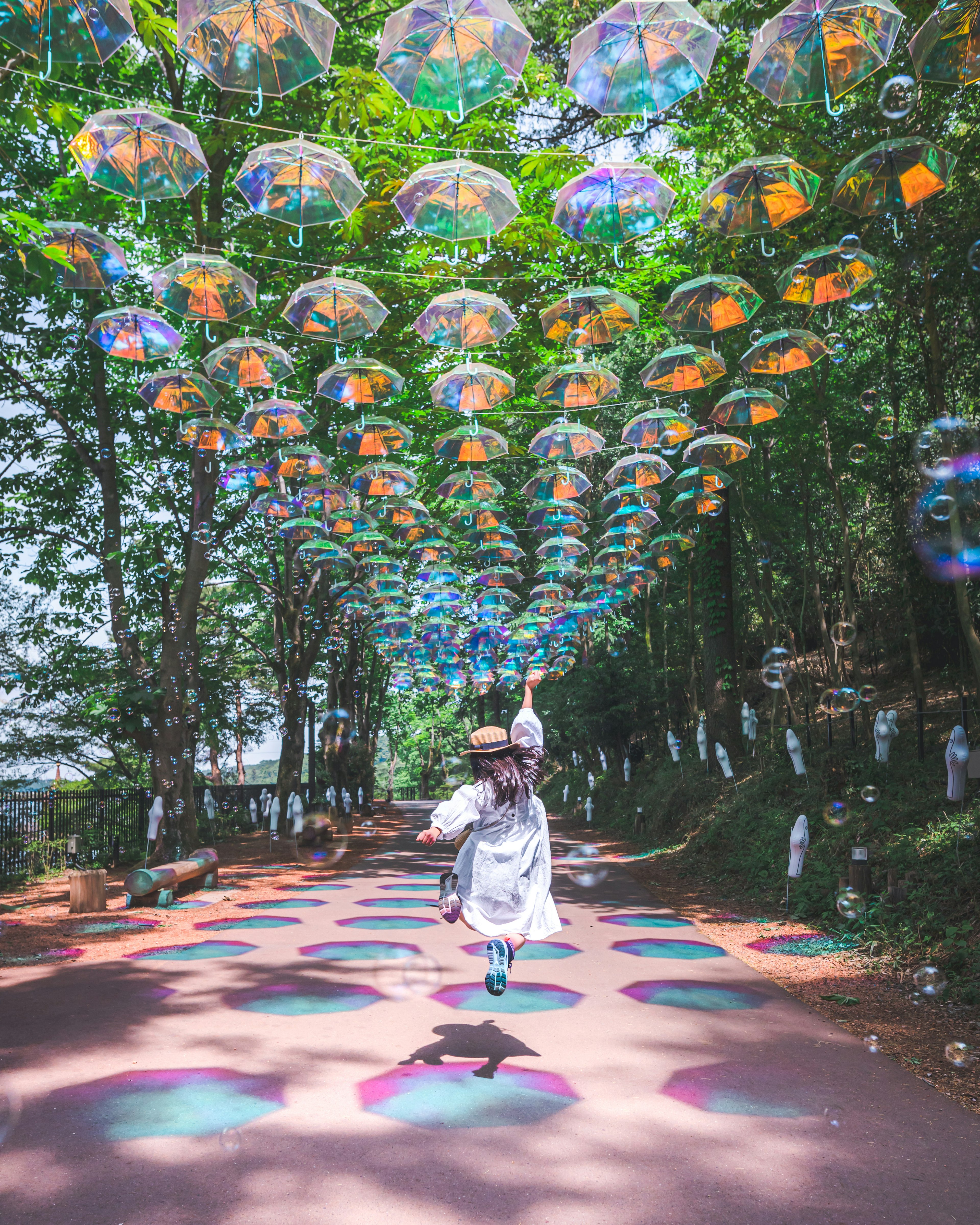 一位女性在郁郁蔥蔥的環境中走在五彩繽紛的道路上，頭頂懸掛著傘