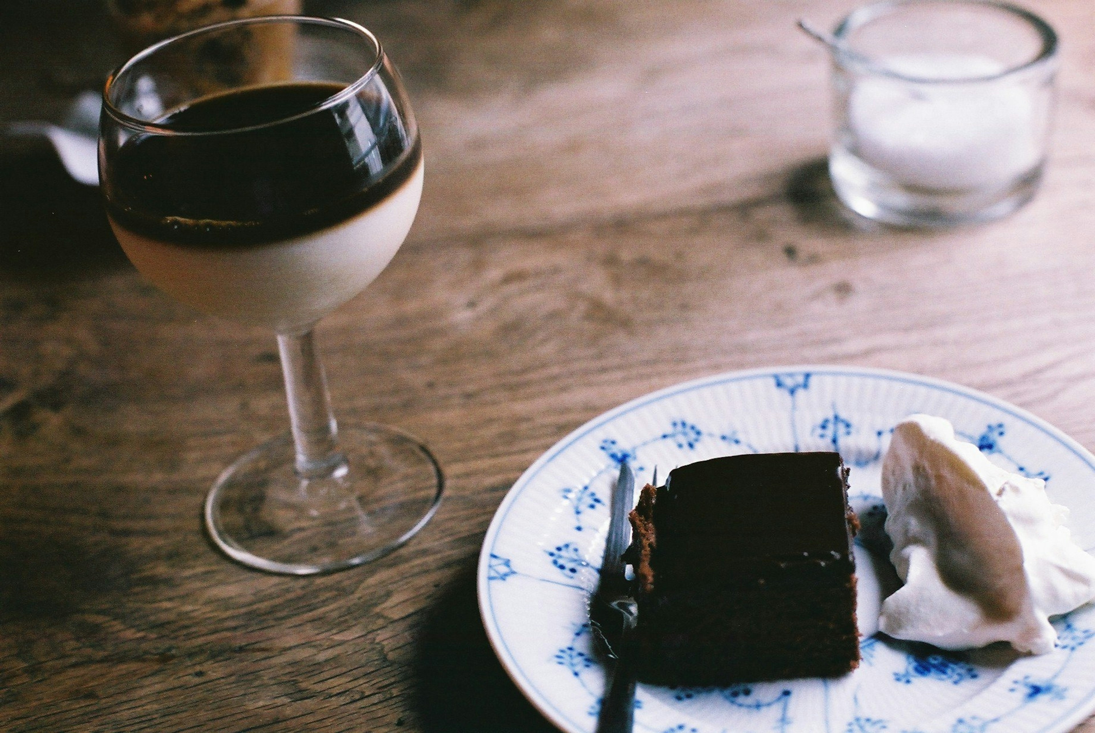 Ein Dessert und ein Getränk auf einem Tisch mit einem Glas Getränk, einem Schokoladenkuchen auf einem blauen Blumenteller und einer Kugel Eis