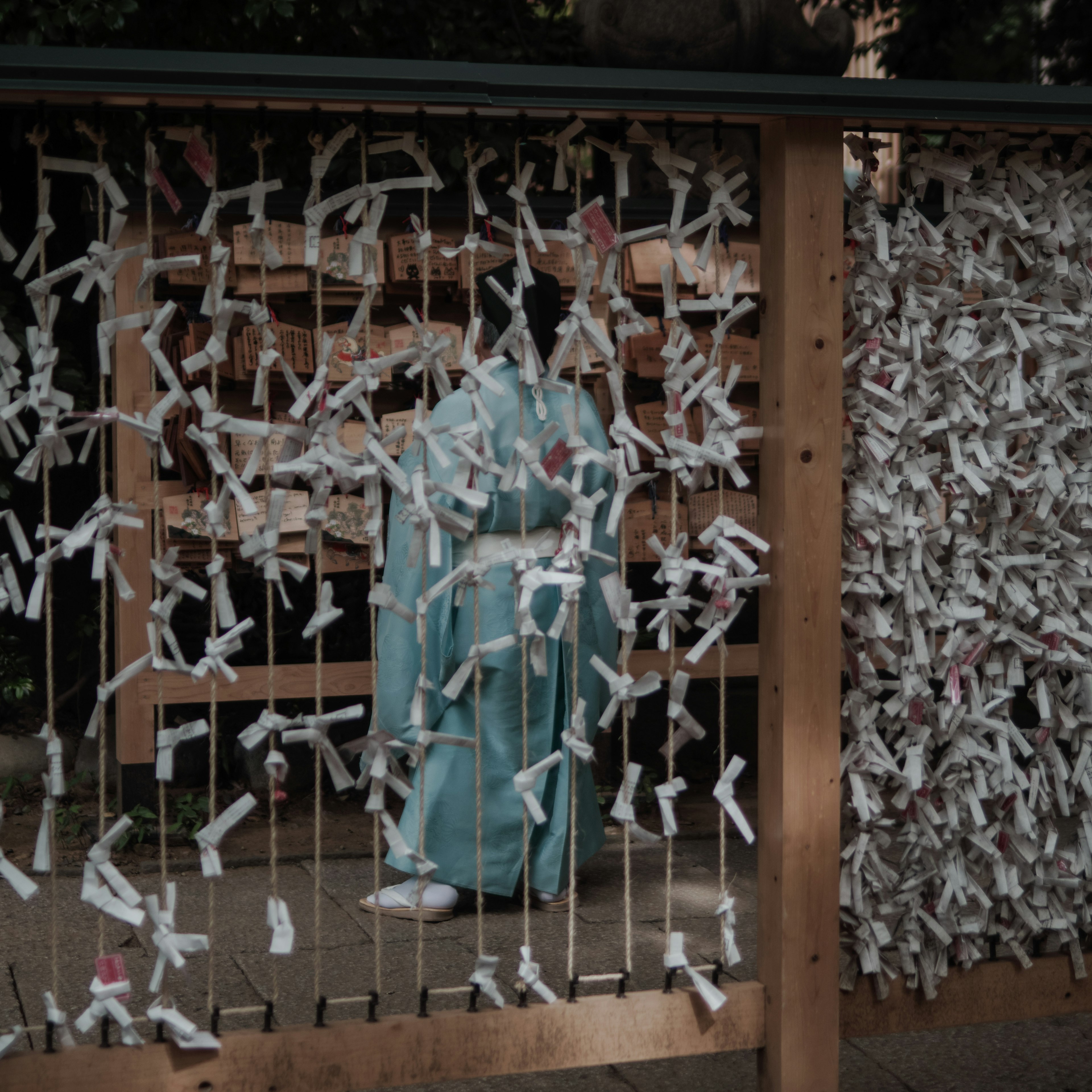 Una donna in kimono blu si trova davanti a una recinzione di legno decorata con strisce bianche di omikuji