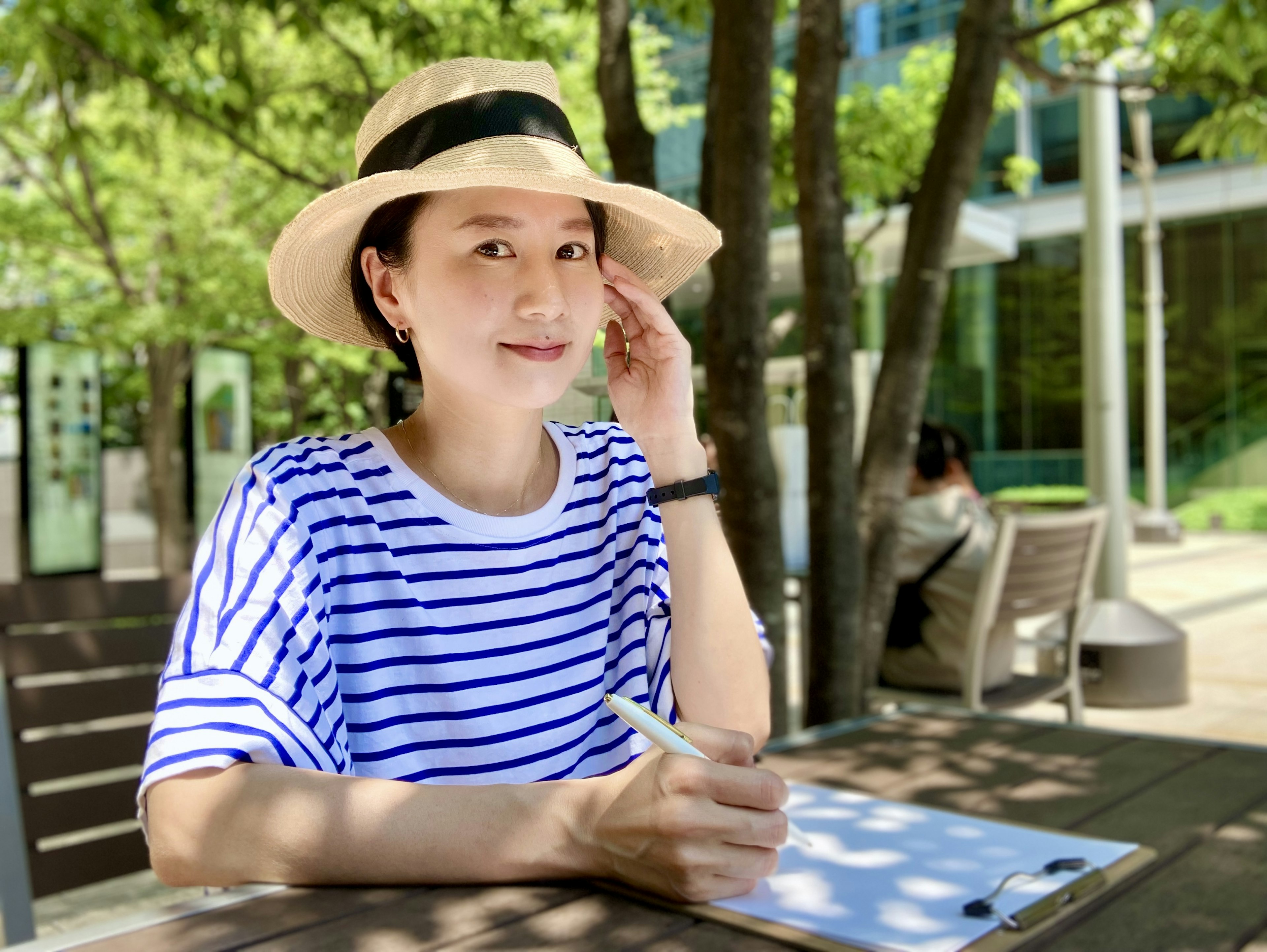 A woman wearing a striped shirt and a hat sitting in the shade taking notes on a notebook