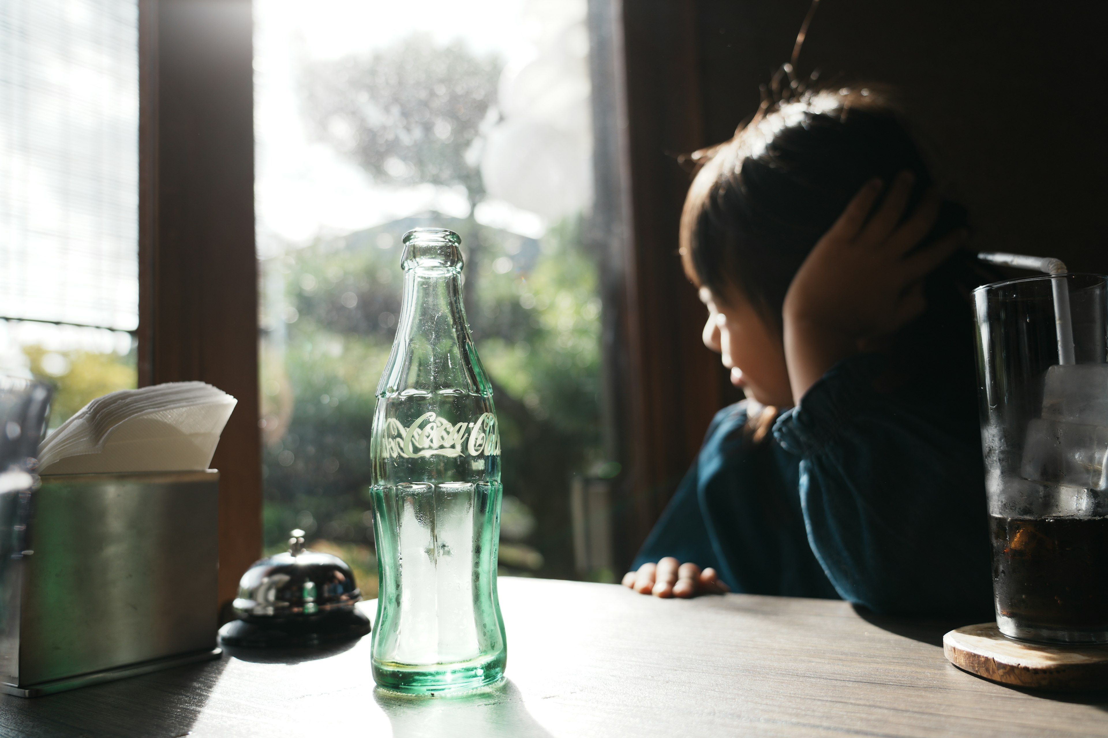 A child looking out the window with a green Coca-Cola bottle on the table