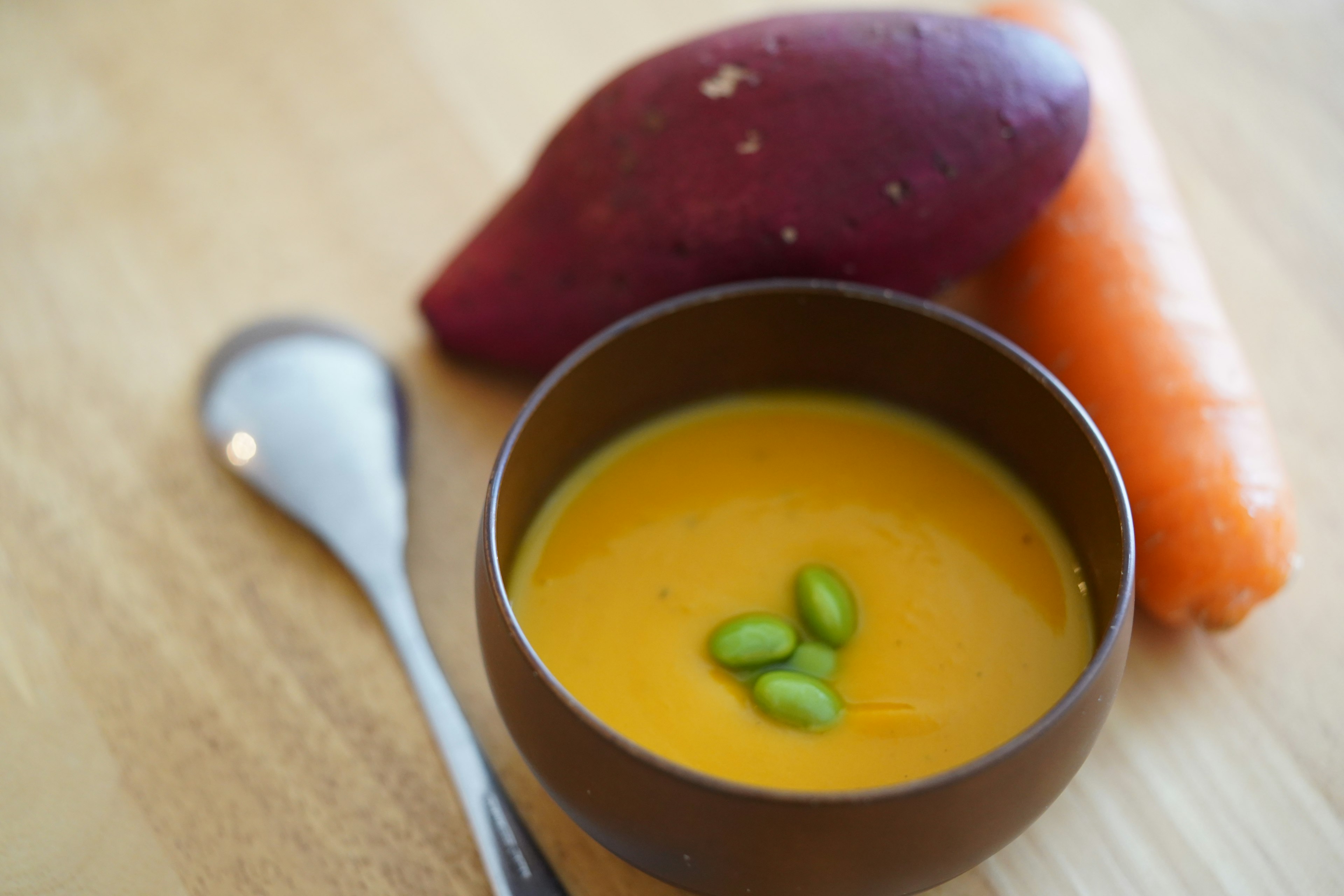 Eine Schüssel mit orangefarbener Suppe, garniert mit grünen Bohnen, umgeben von Süßkartoffel und Karotte