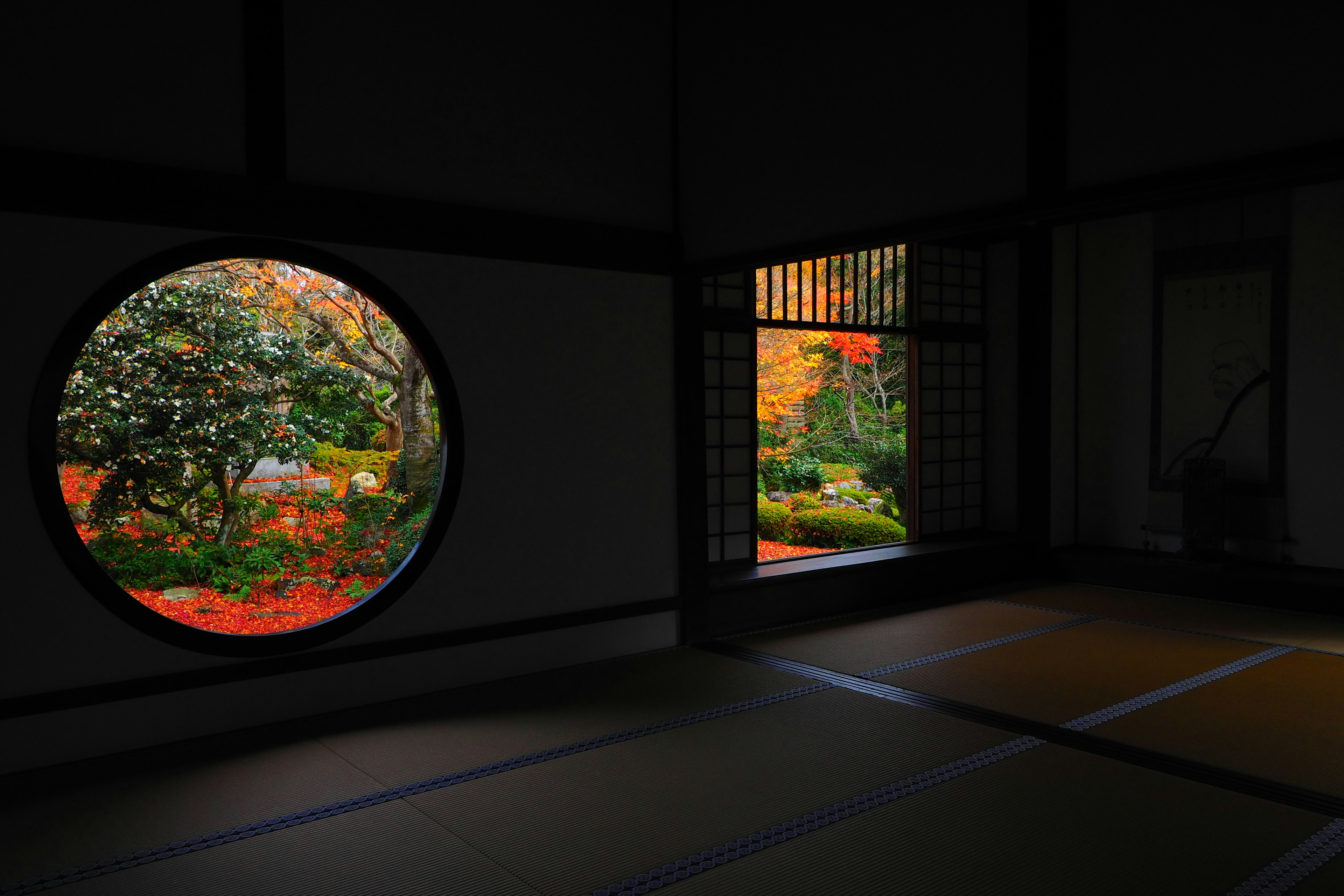 Habitación oscura con ventanas circulares y de rejilla que muestran un paisaje otoñal vibrante