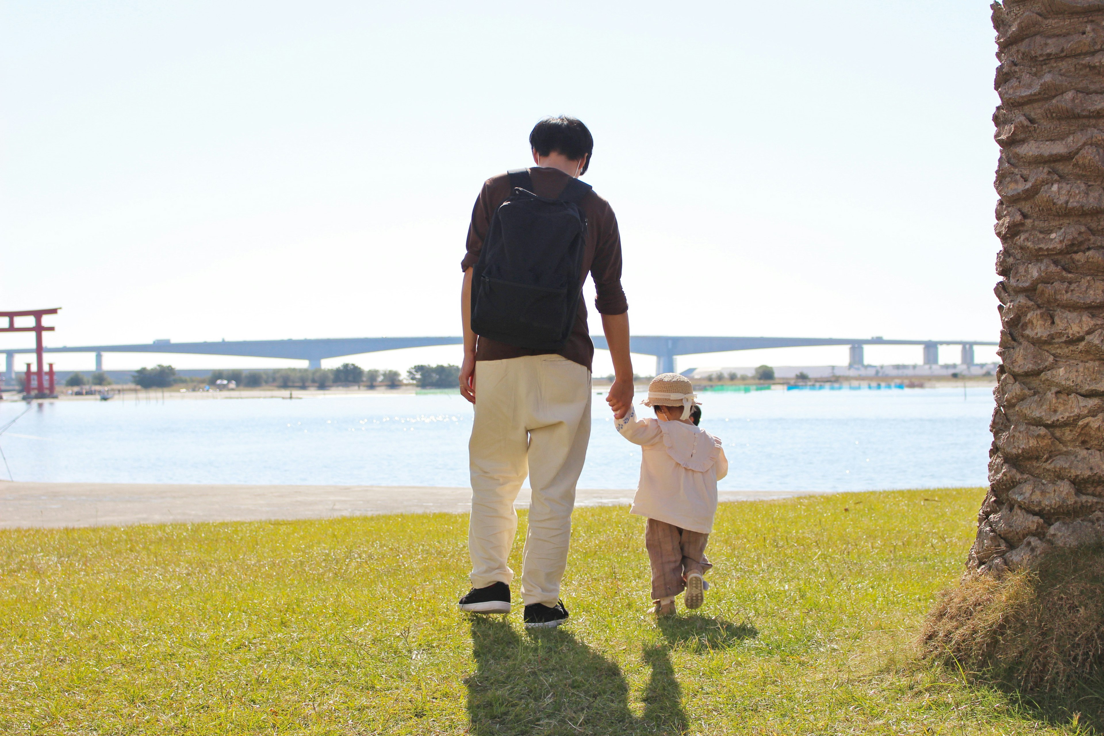 A father and child walking hand in hand by the river with a bridge in the background