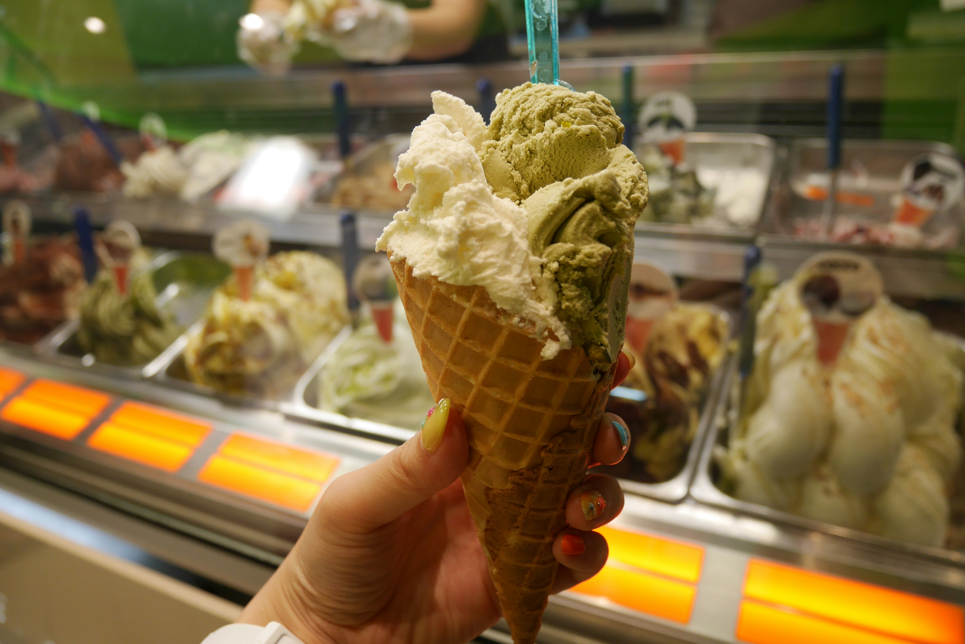 Mano sosteniendo un cono de waffle con tres bolas de helado frente a un exhibidor de helados