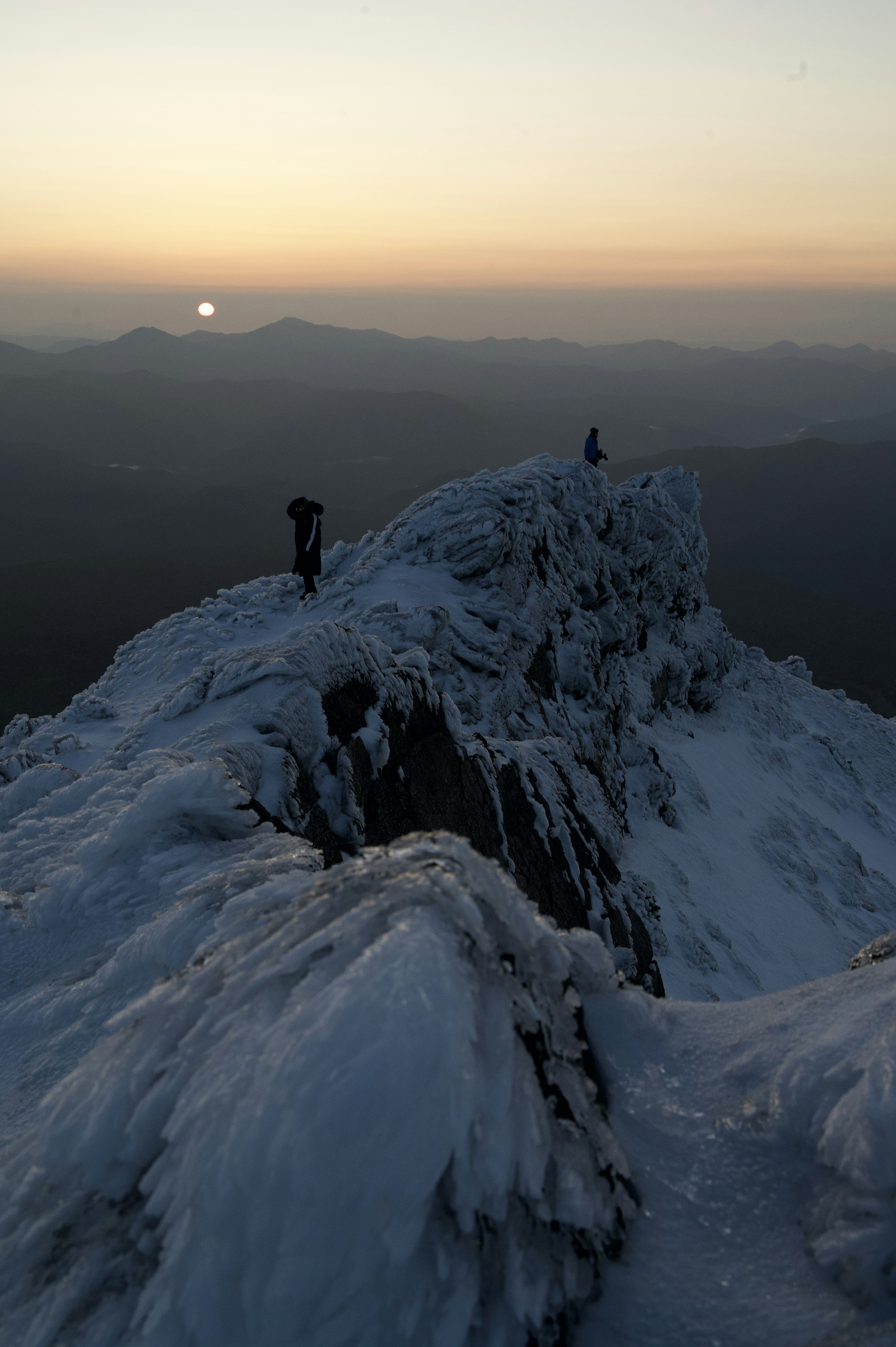 雪に覆われた山頂で二人の登山者が夕日を背景に立っている