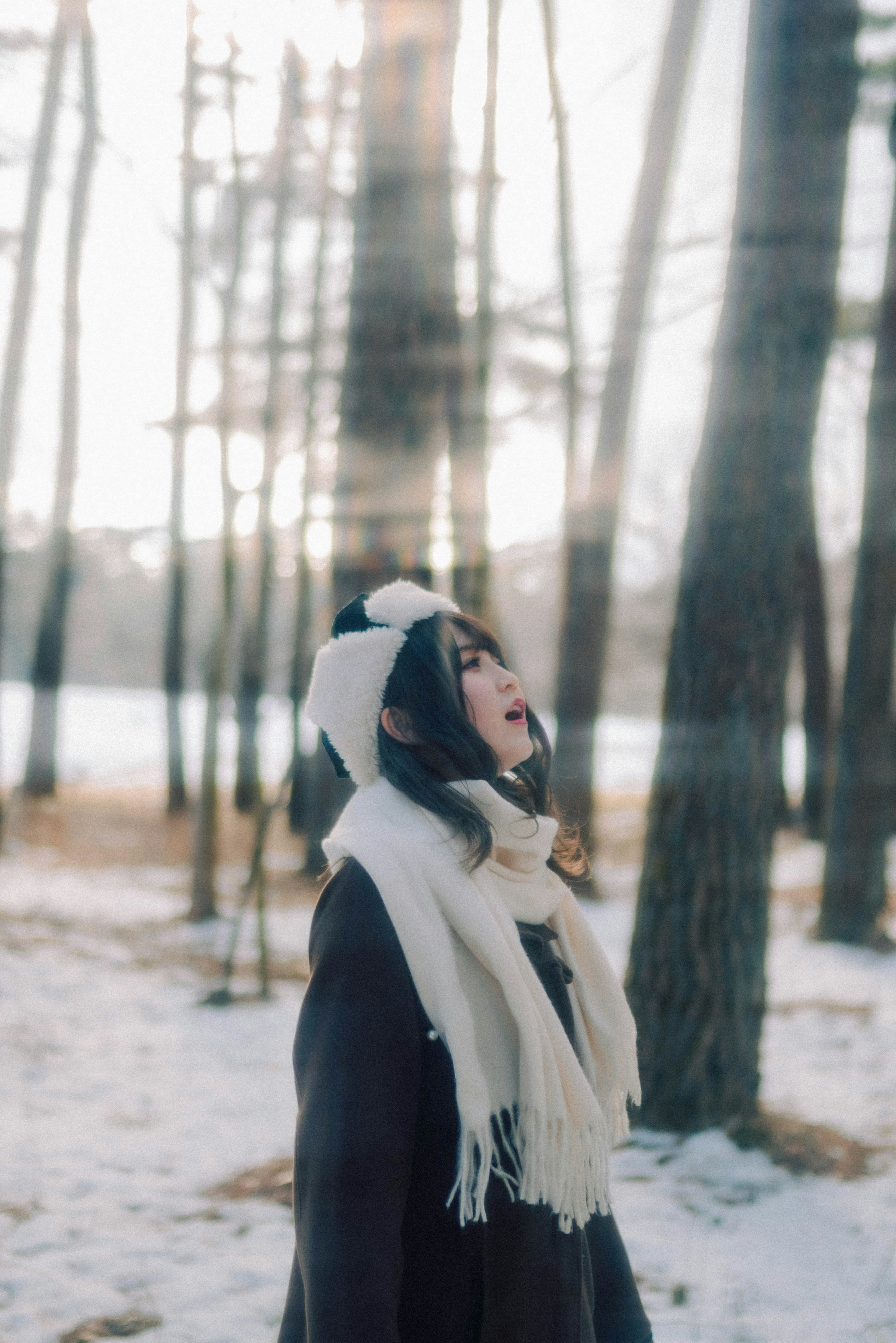 Eine Frau schaut in einem verschneiten Wald nach oben und trägt Wintermode mit Schal und Mütze