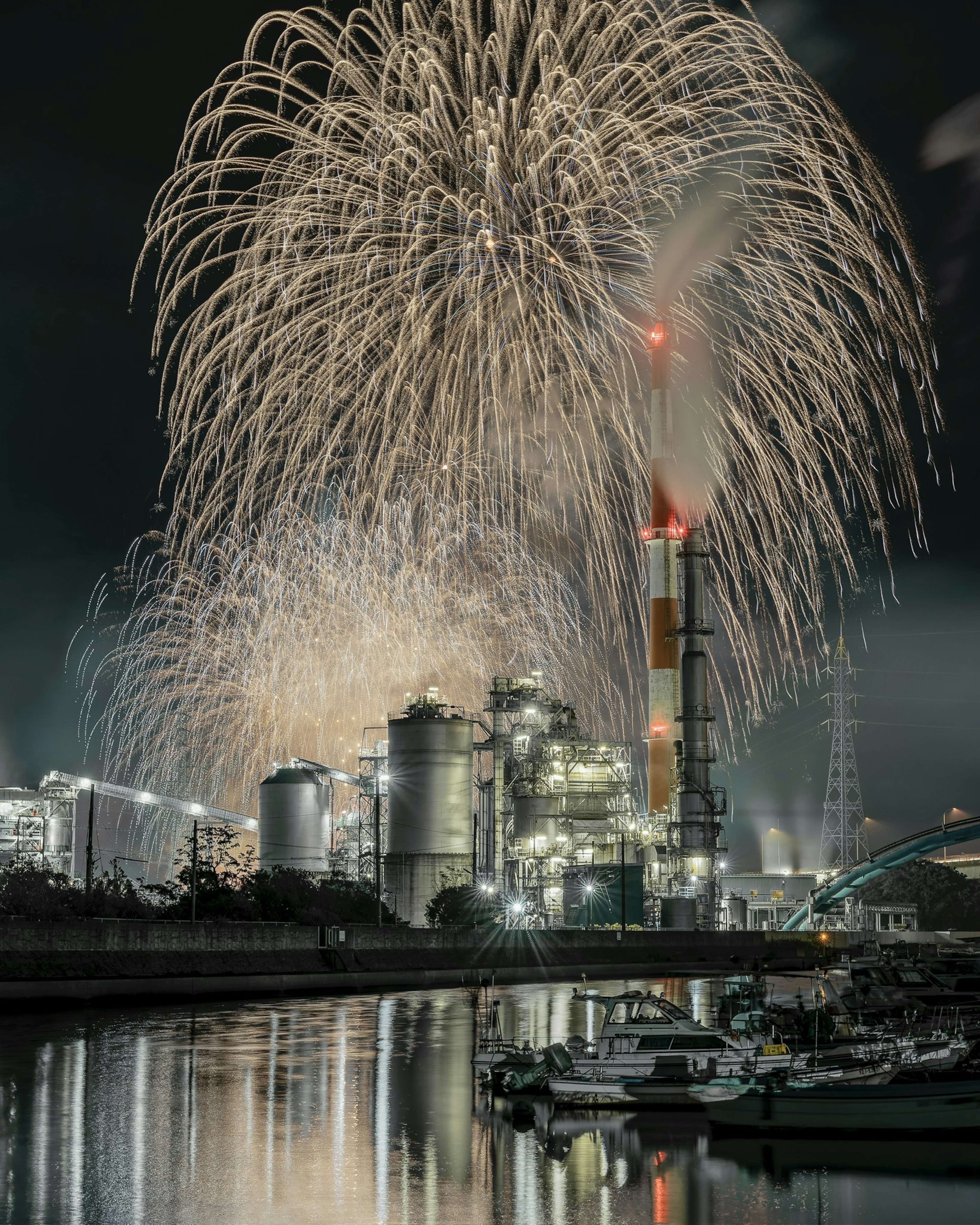 Los fuegos artificiales iluminan el cielo nocturno sobre una fábrica con chimeneas reflejadas en el agua