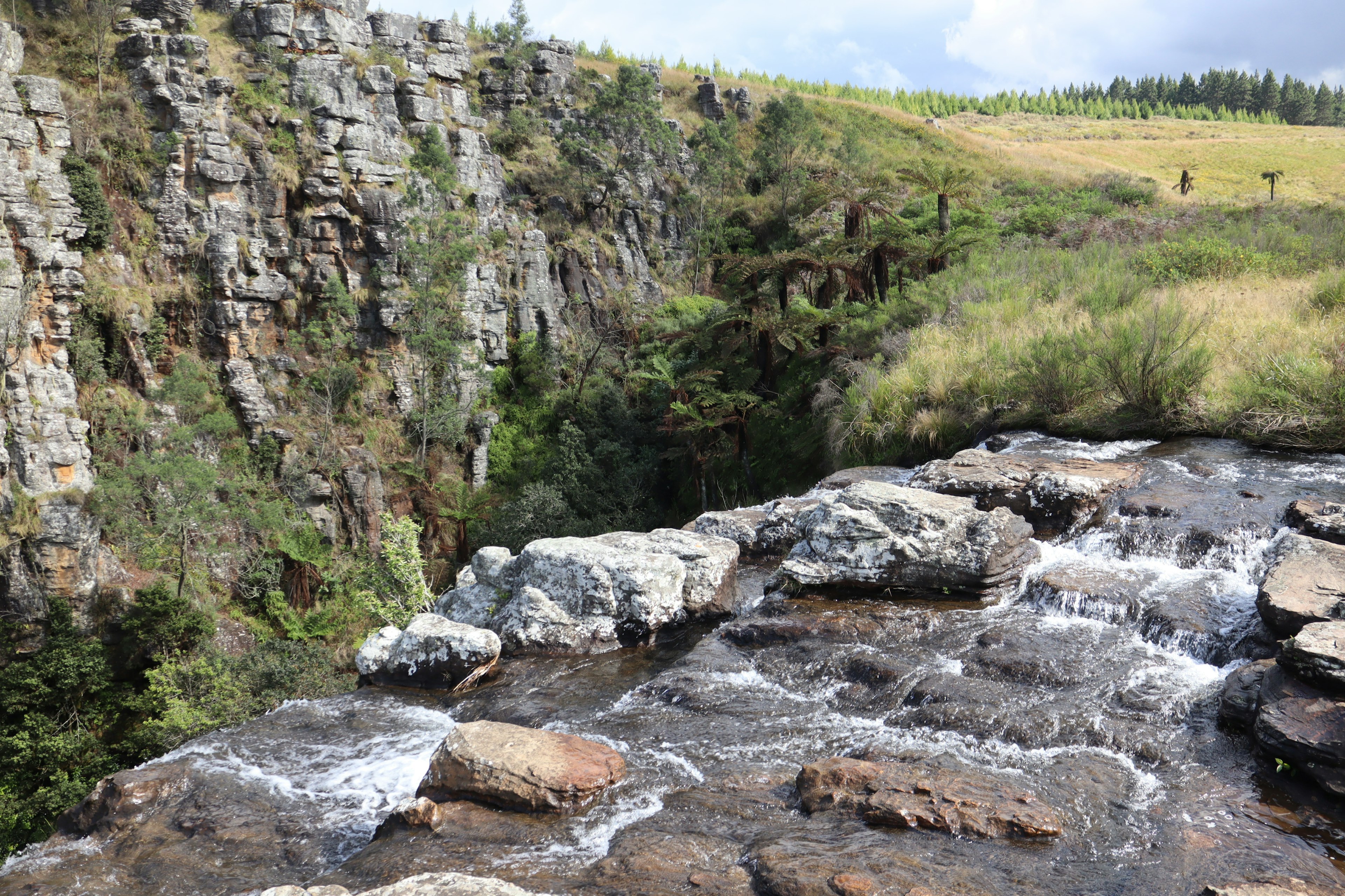 Vista panoramica di una cascata che scorre su rocce con una scogliera e prati verdi sullo sfondo