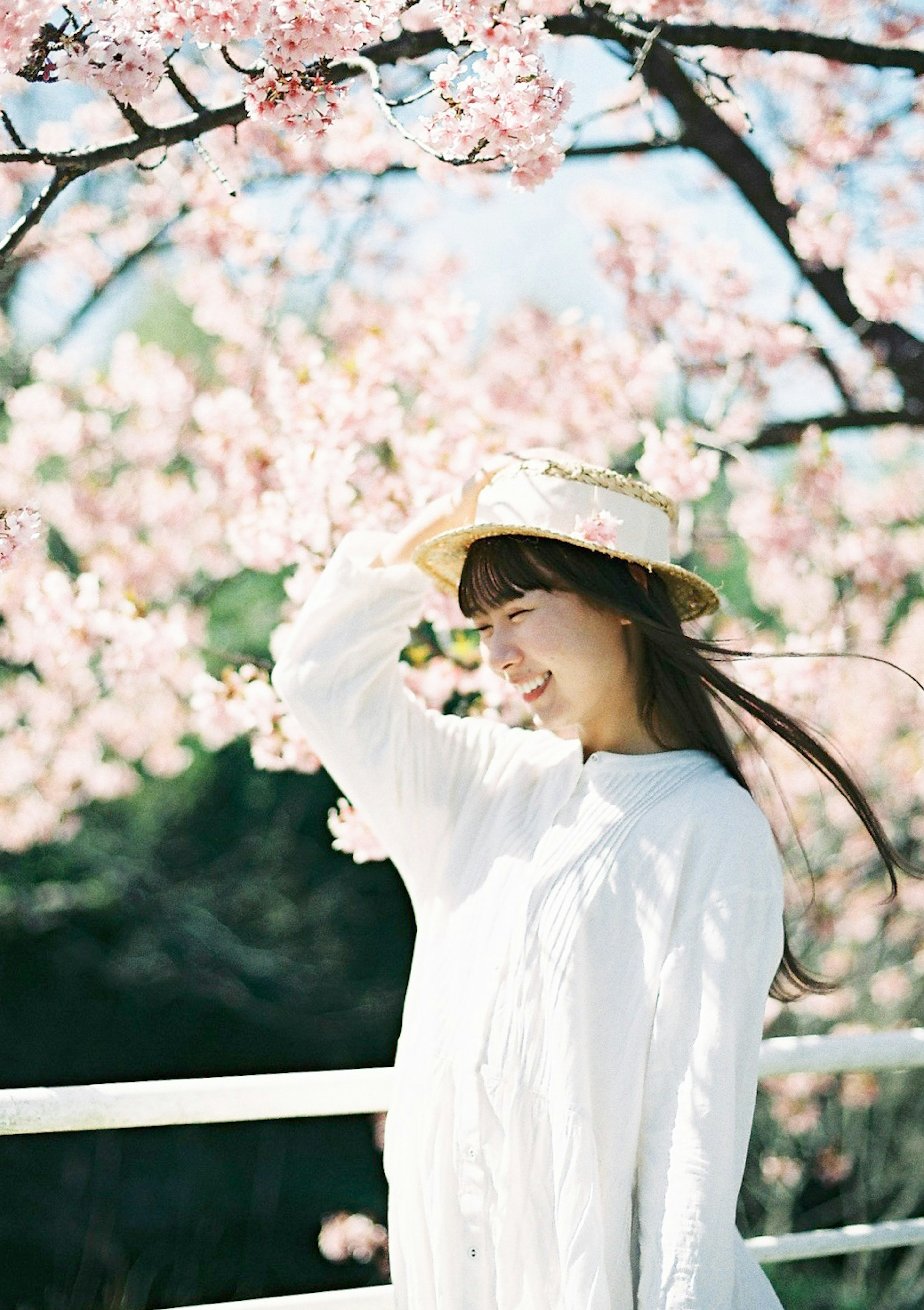 Una mujer con vestido blanco y sombrero sonriendo debajo de un árbol de cerezo