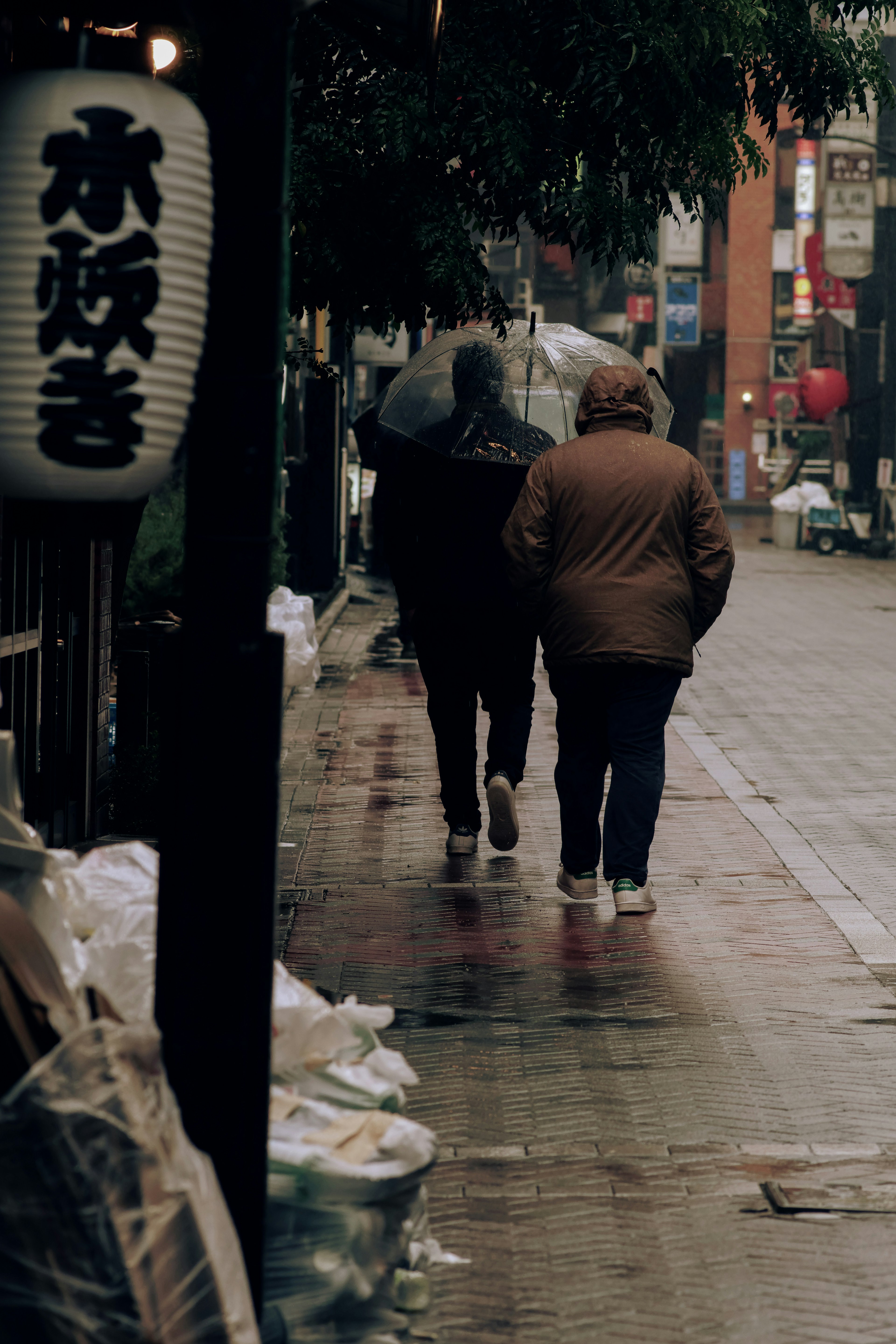 Two figures walking in the rain with a Japanese sign nearby