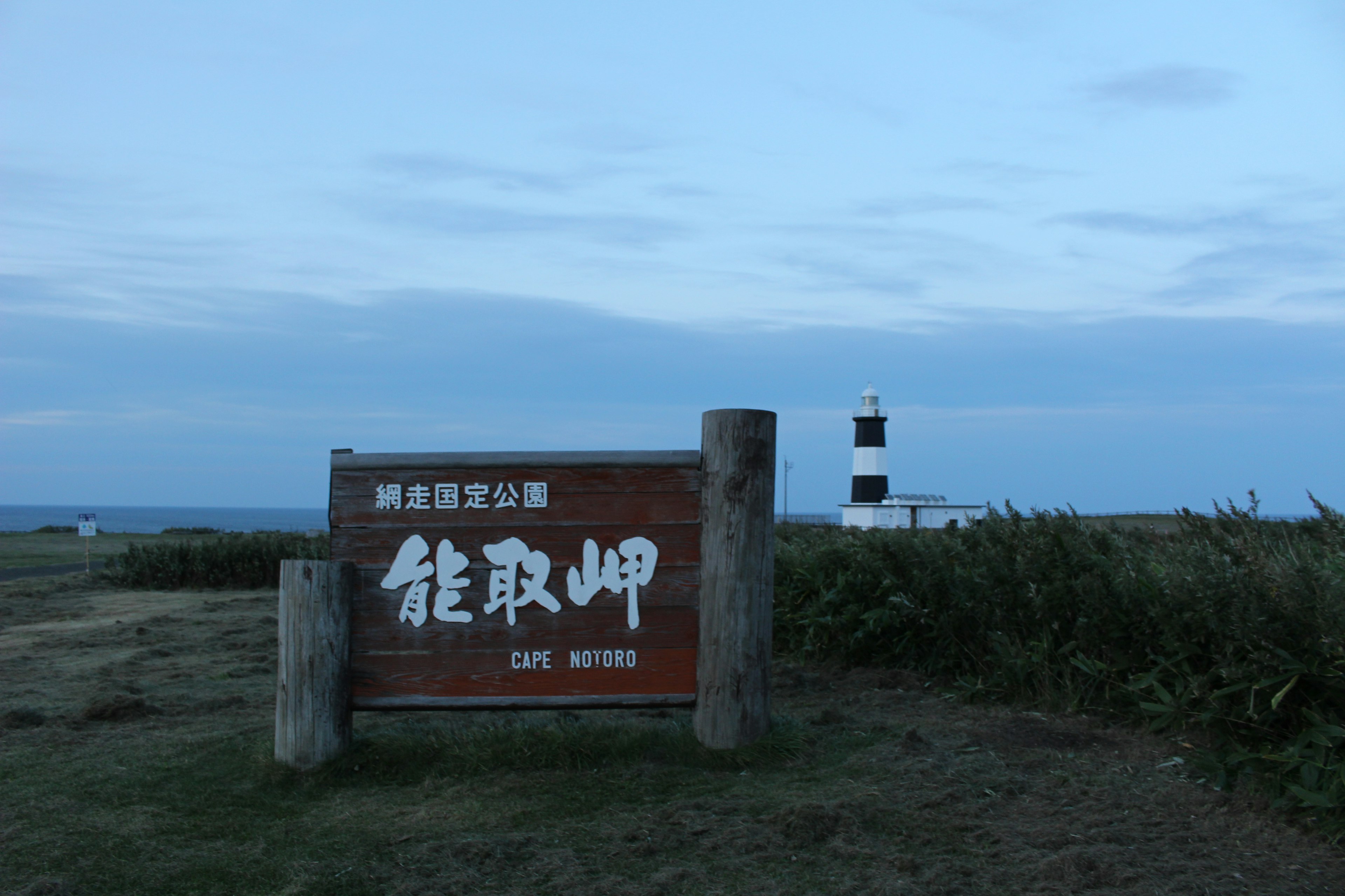 Schild für Notoro Misaki mit Leuchtturm im Hintergrund blauer Himmel und Grasfläche