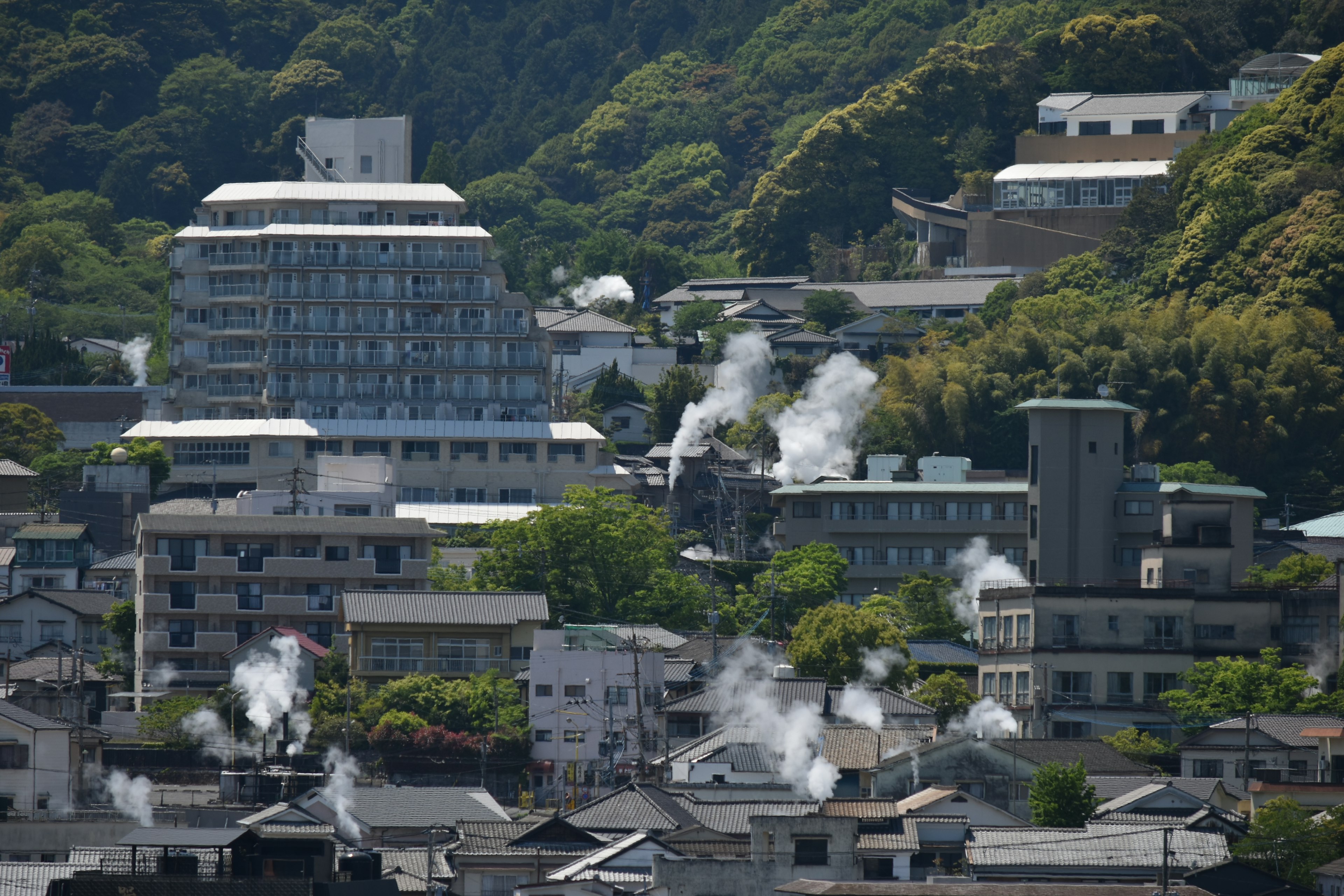 温泉街の建物と緑豊かな山々が見える風景で、湯気が立ち上る