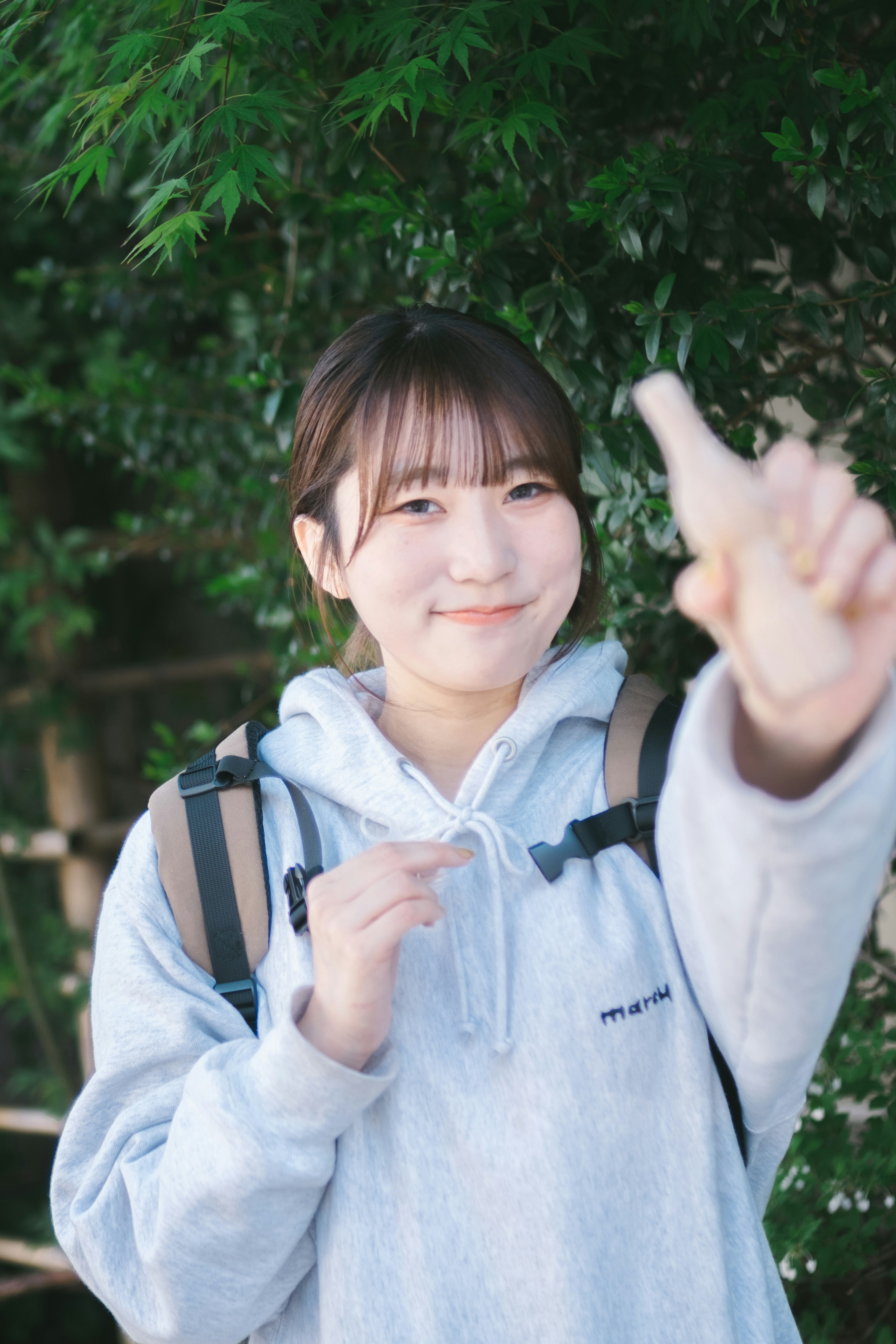 Young woman in gray hoodie smiling and making a peace sign with green plants in the background