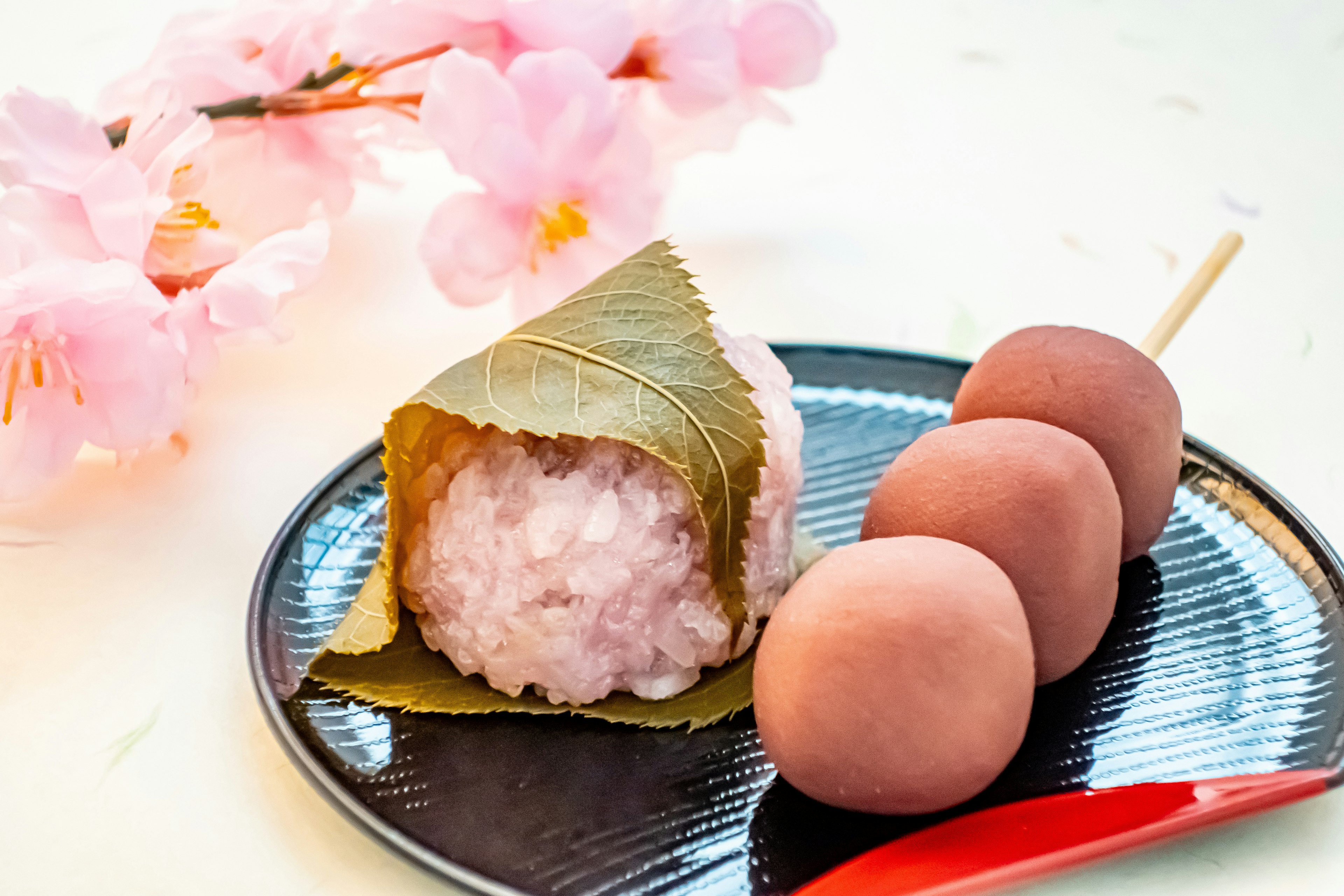 Bola nasi yang dibungkus daun di samping bunga sakura dan tiga dango di atas piring