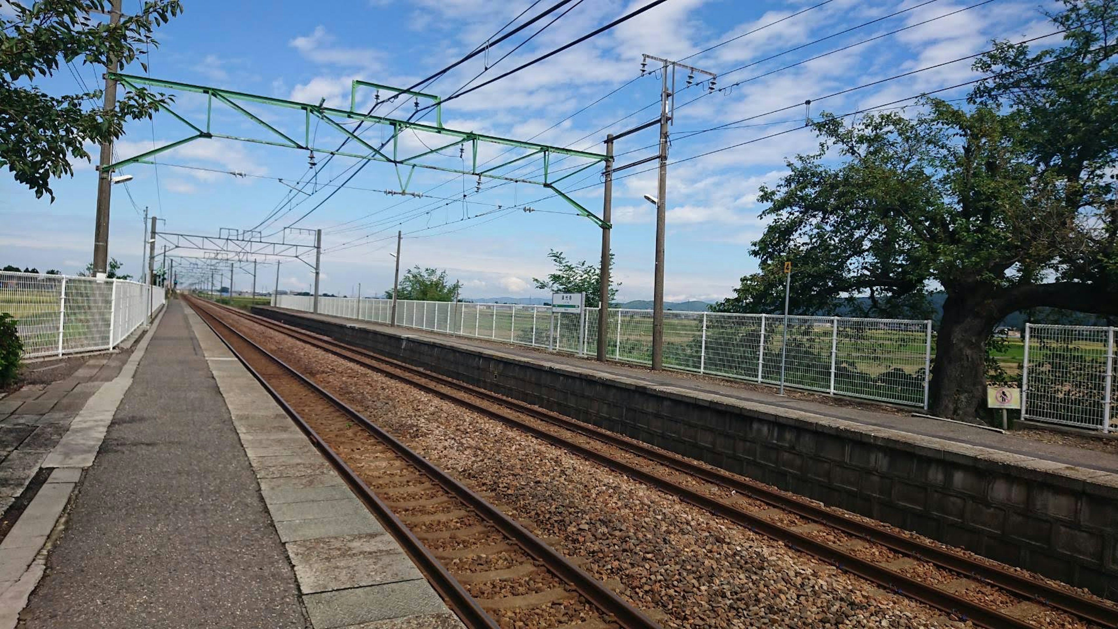 Bahnhofansicht mit Gleisen und Zaun blauer Himmel und grüne Bäume sichtbar