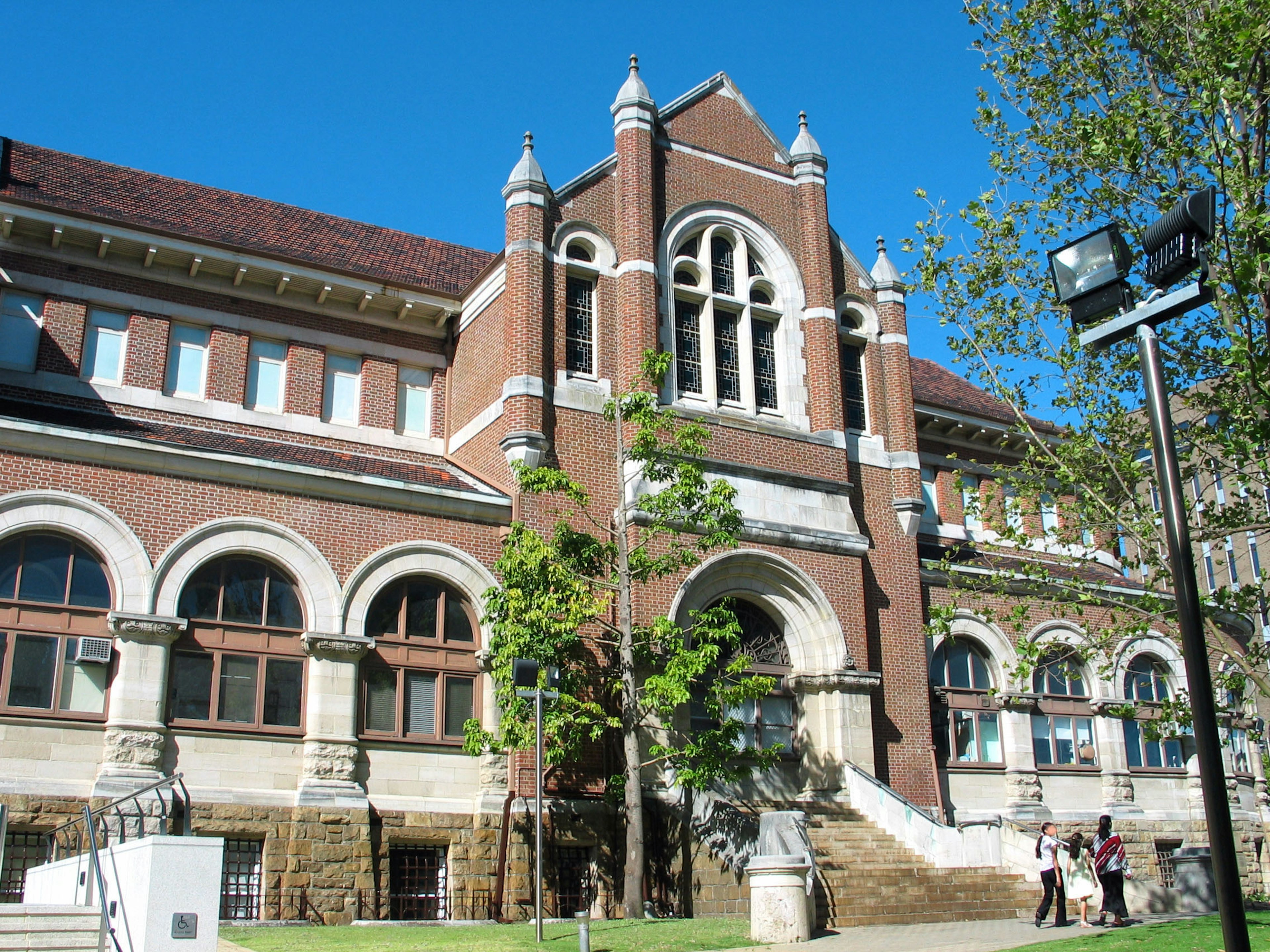 Historic building exterior with reddish brick, Gothic-style windows, green trees, clear blue sky