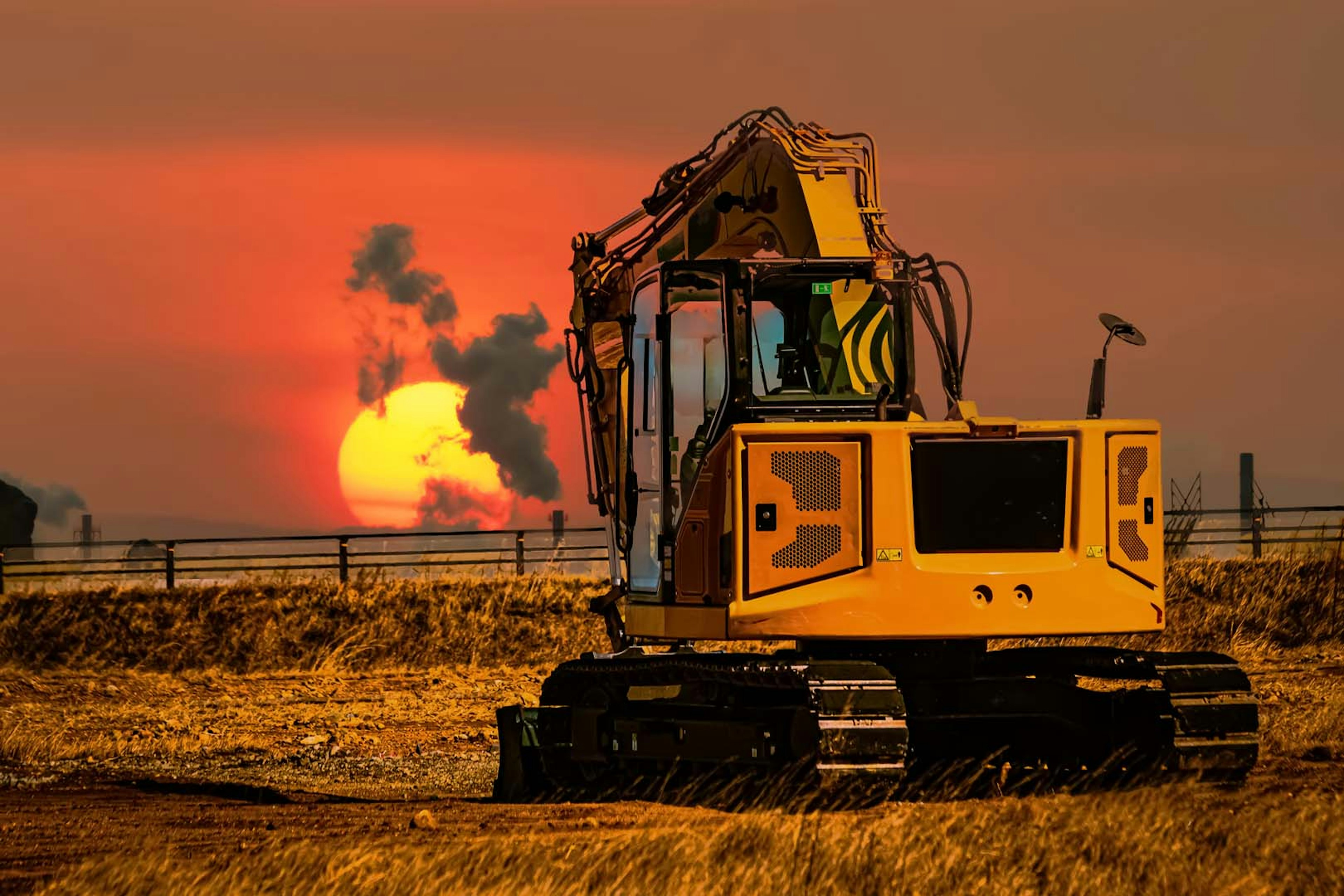 Ekskavator kuning di latar belakang matahari terbenam dengan asap